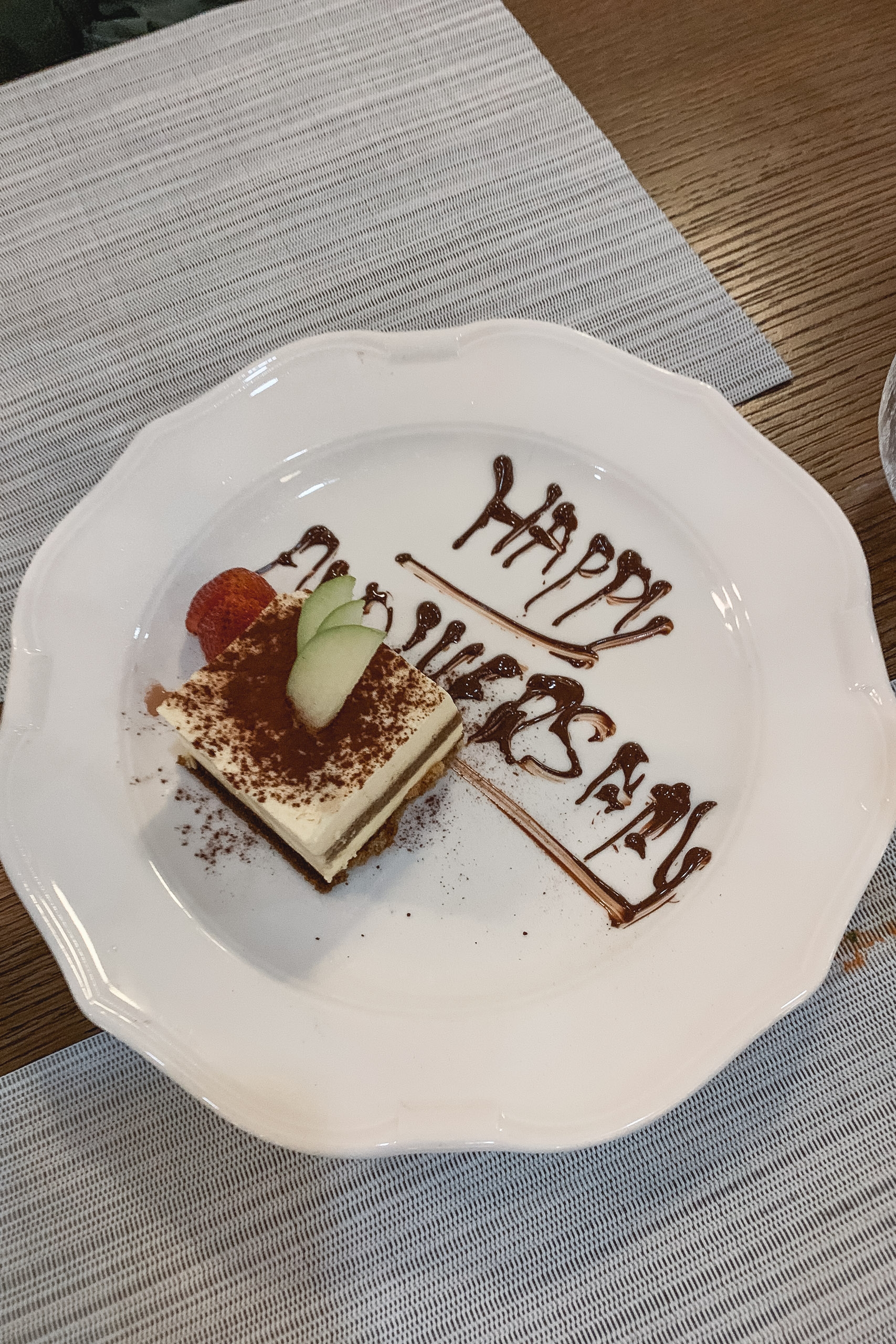 A white scalloped plate displays a layered tiramisu dessert with cocoa powder dusting, topped with fresh strawberry and cucumber slice, accompanied by chocolate script reading "Happy Birthday" on wooden table