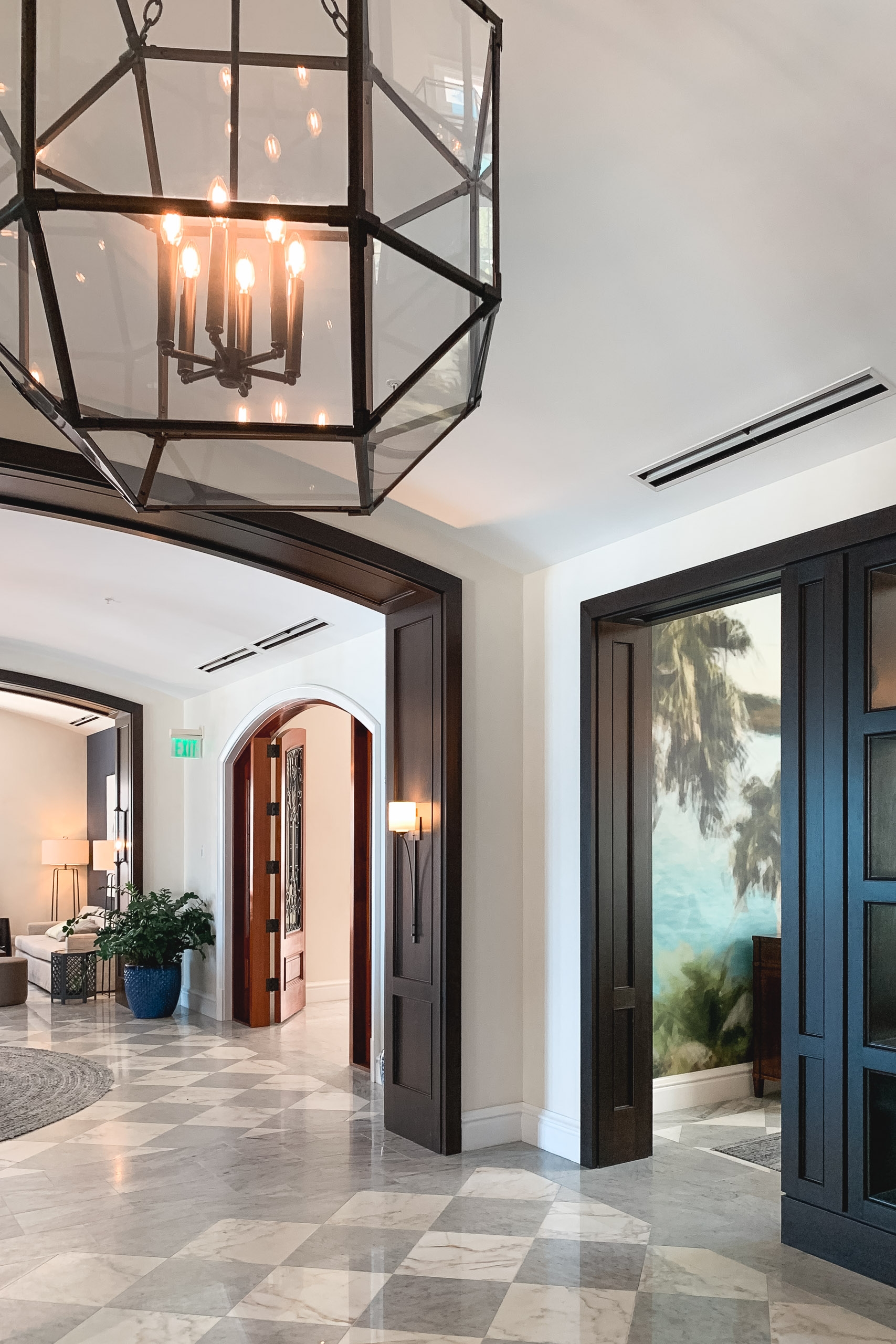 A modern foyer features a geometric black pendant light with glowing candles, polished stone flooring, and dark-trimmed doorways opening to adjacent rooms bathed in natural light