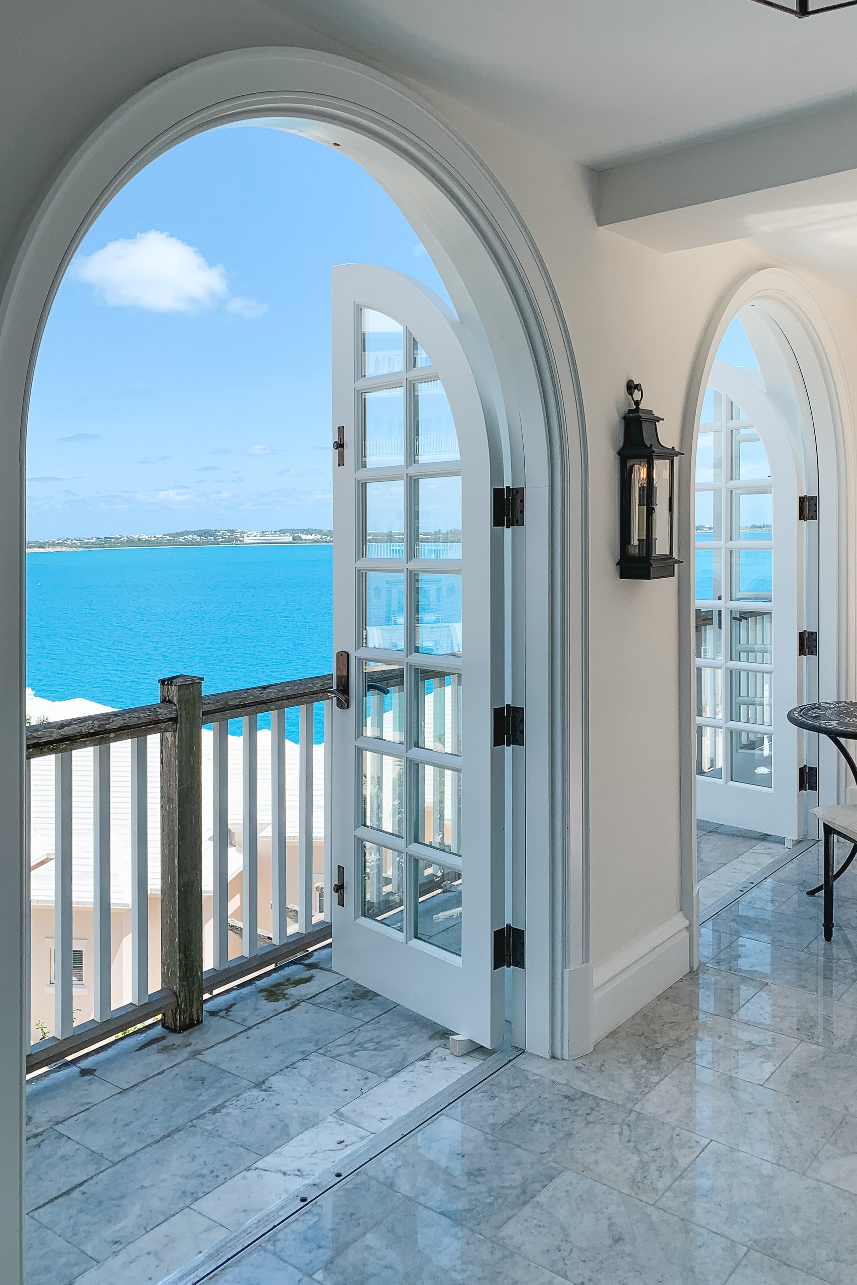 Arched white doors frame a turquoise ocean view from a bright coastal interior, with a black lantern and white railing visible on the sunny balcony beyond