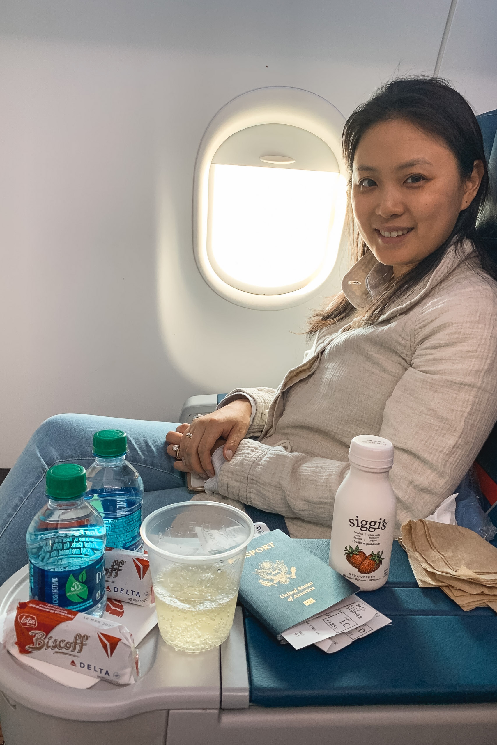 A woman sits in an airplane seat beside a bright window, smiling at the camera while surrounded by a tray table of beverages, snacks, and travel documents in warm cabin lighting