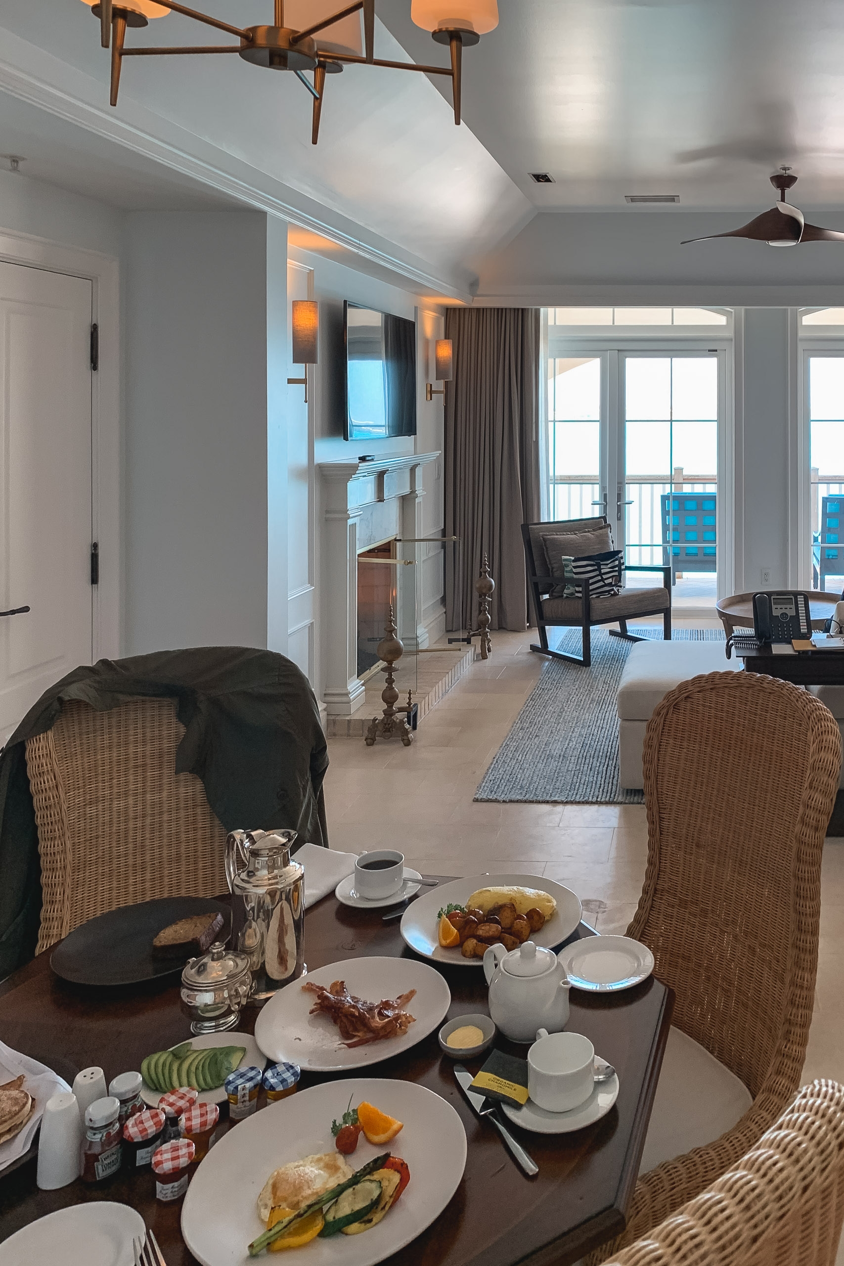 A coastal hotel suite features a dining table set for breakfast with woven chairs, ocean views through glass doors, and mid-century modern ceiling fixtures illuminating the bright, contemporary space