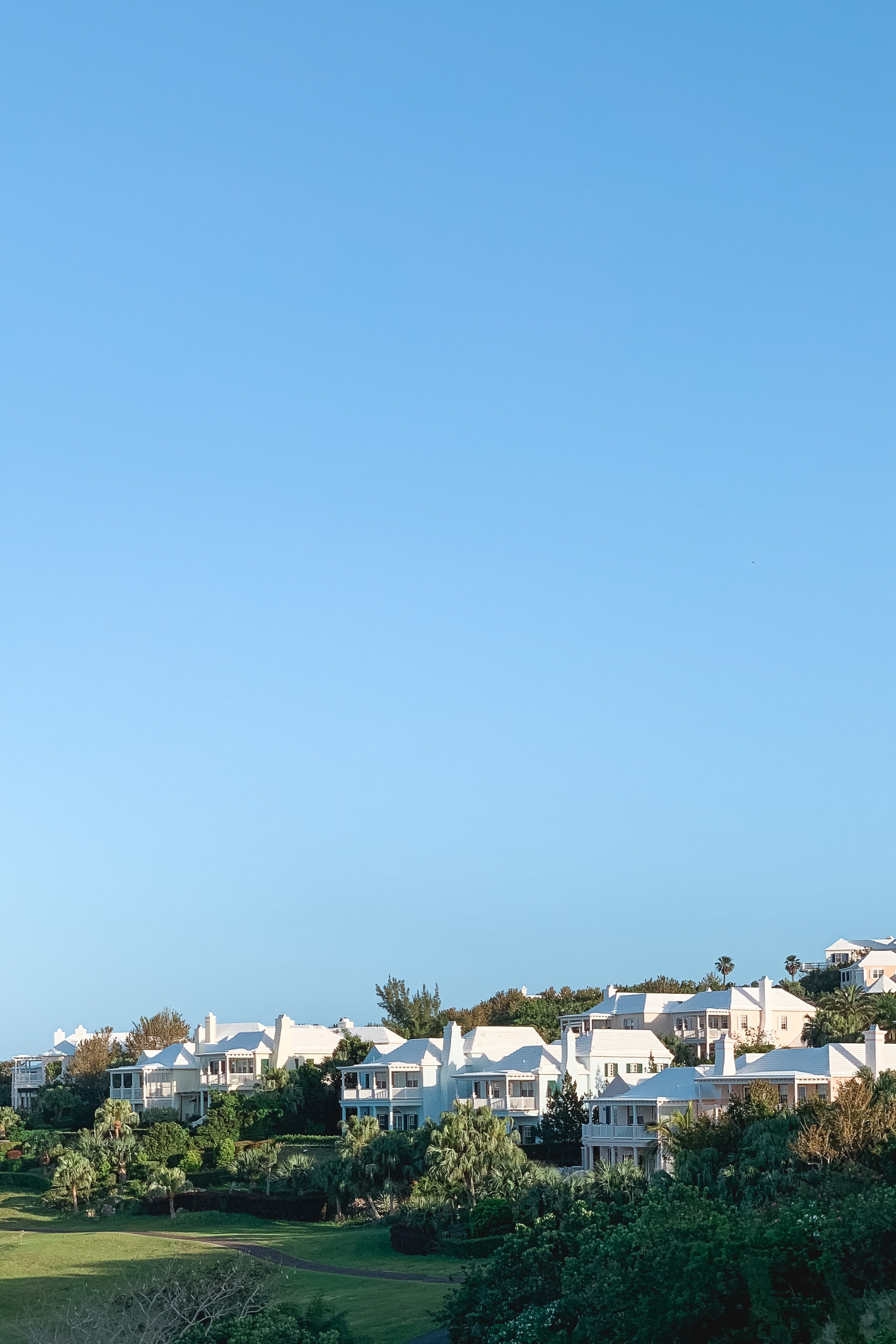 White colonial-style houses with blue roofs nestle among lush green vegetation under a clear blue sky in Bermuda's coastal residential area