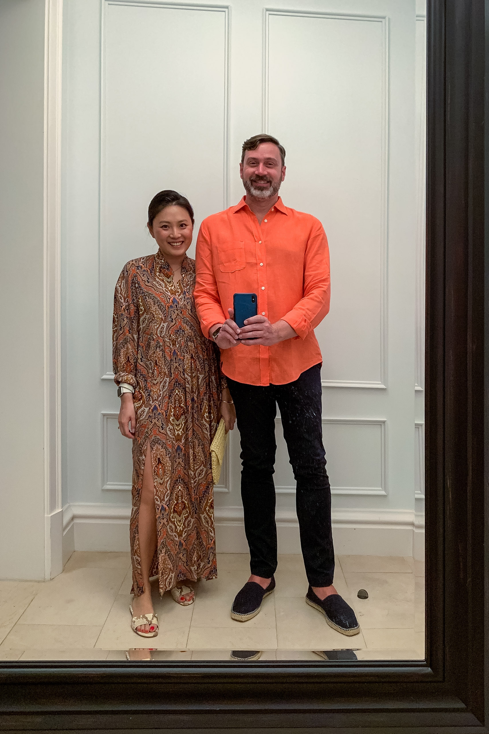 A man in a bright orange shirt holding a phone and a woman in a patterned bronze dress stand together against white paneled walls in an elegant interior space