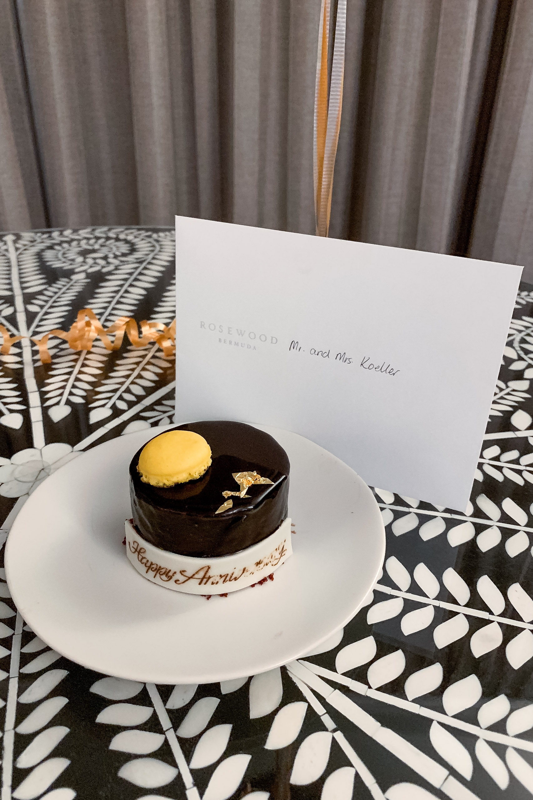 An elegant dessert plate holds a two-toned cake with dark chocolate top and white base, topped with a golden dome, positioned beside a white invitation card on a geometric patterned table with gray curtains behind