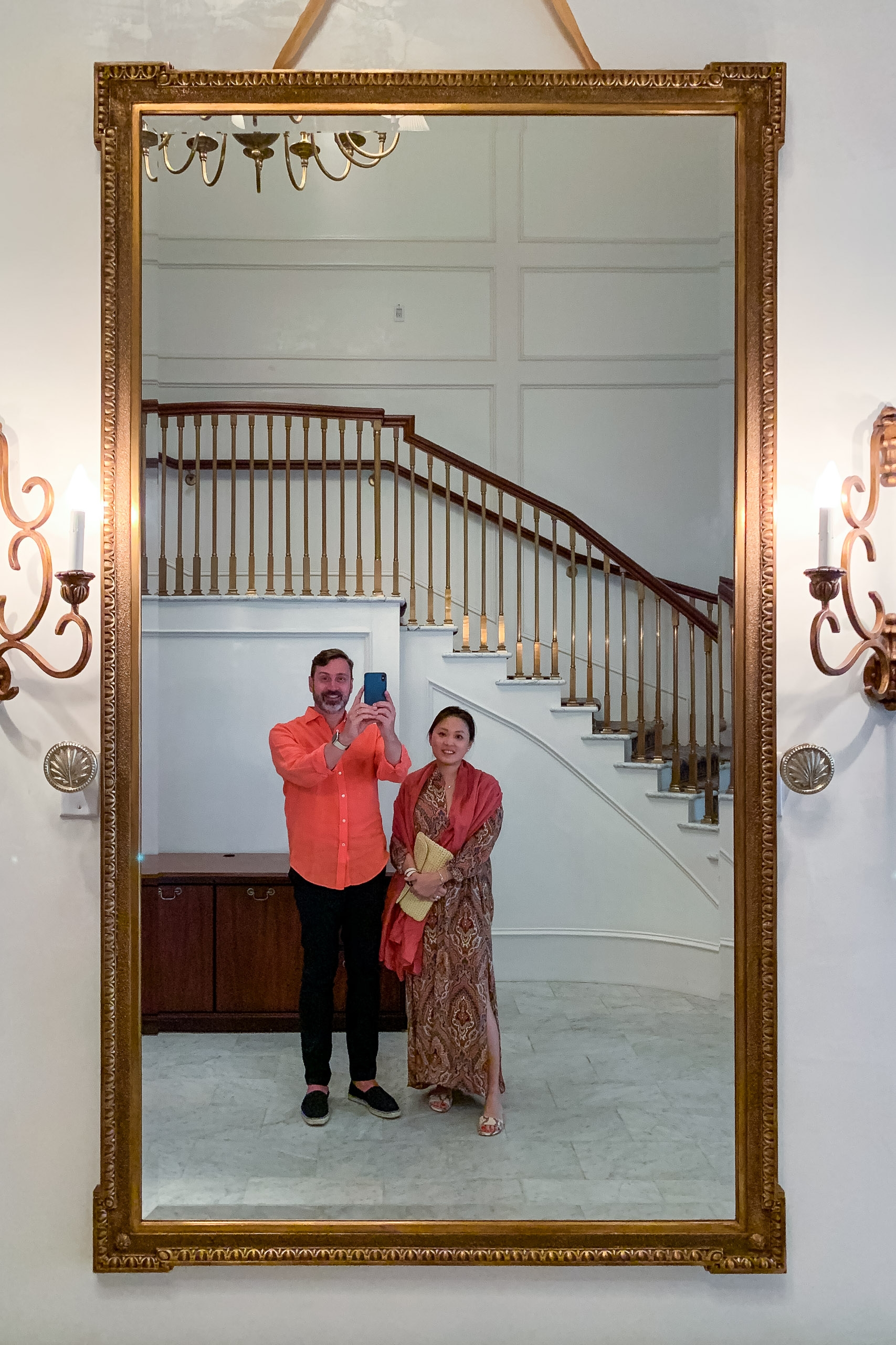 Two people in formal red attire stand in an ornate gold-framed mirror reflecting an elegant staircase and classical interior hallway behind them