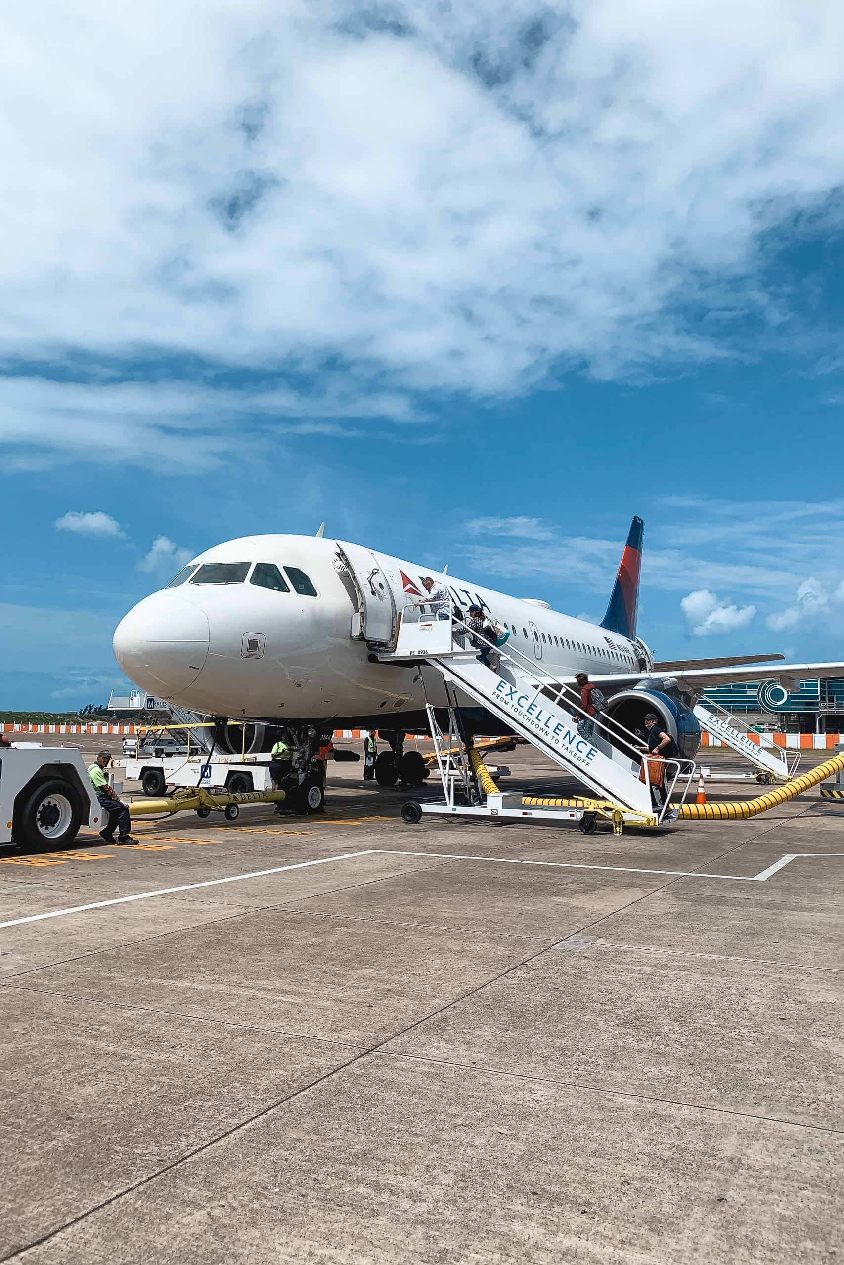 A white commercial aircraft with red tail markings sits parked at an airport gate with boarding stairs attached, under a partly cloudy blue sky