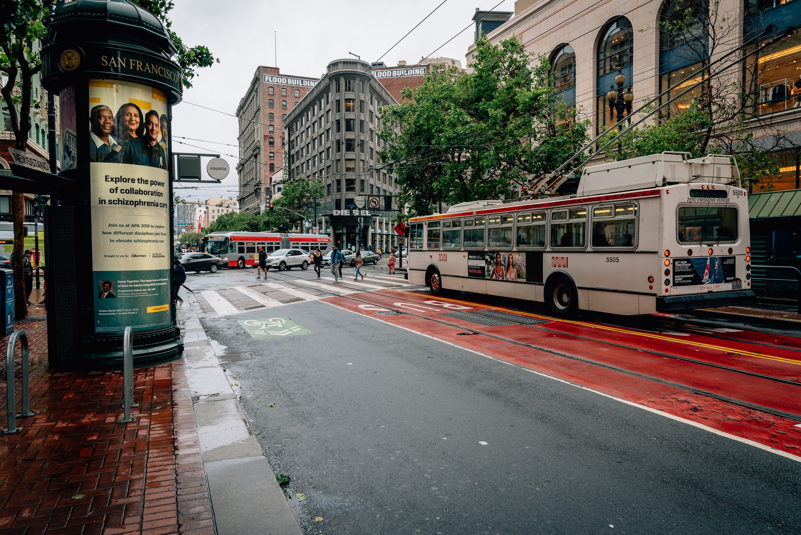 A cream-colored cable car travels along San Francisco's Market Street past historic architecture, with red track lines, green foliage, and an informational kiosk in the foreground under overcast skies.