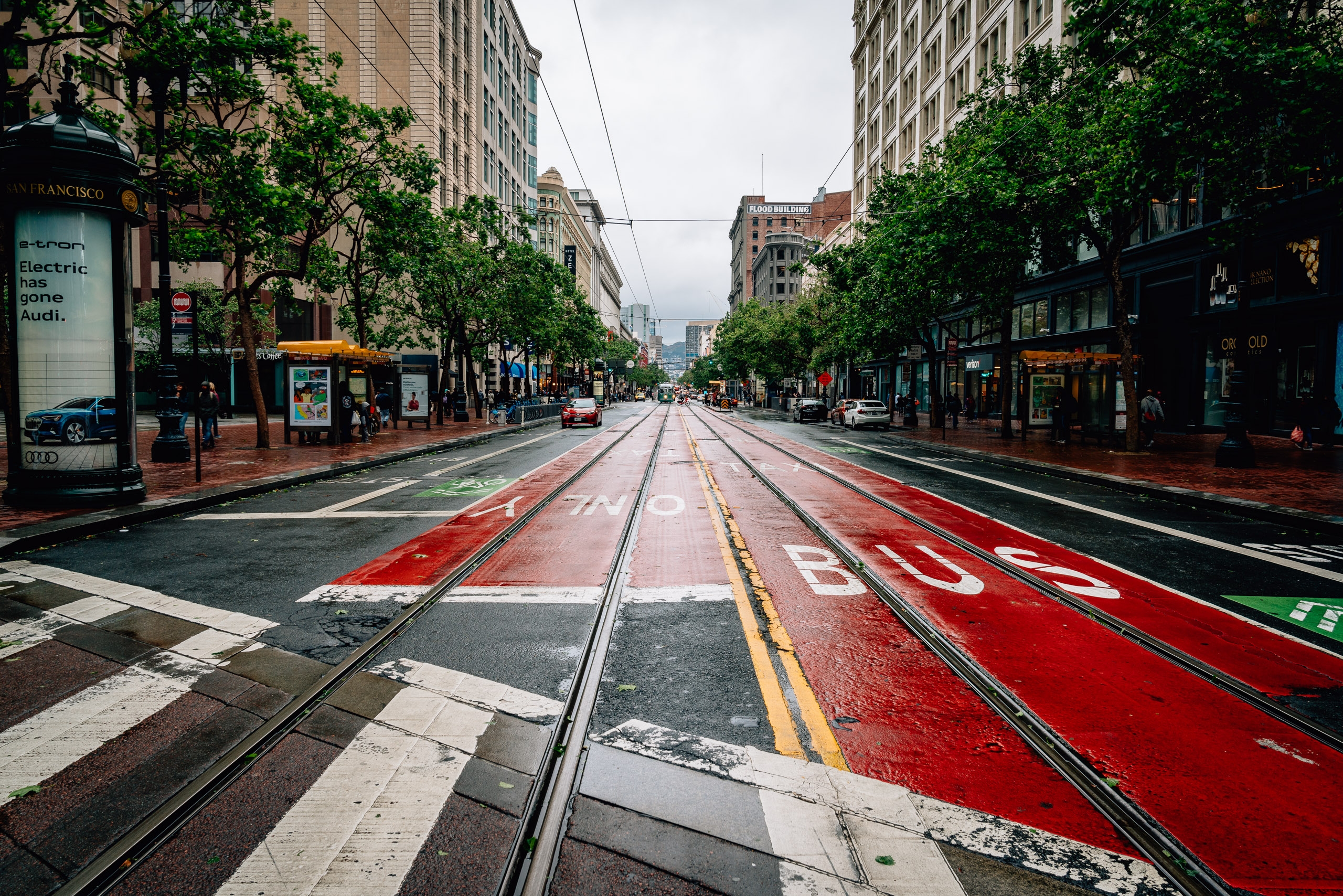 Market Street's cable car tracks recede into the distance between tall downtown buildings and mature trees, with red bus lane markings and storefronts visible under overcast San Francisco light