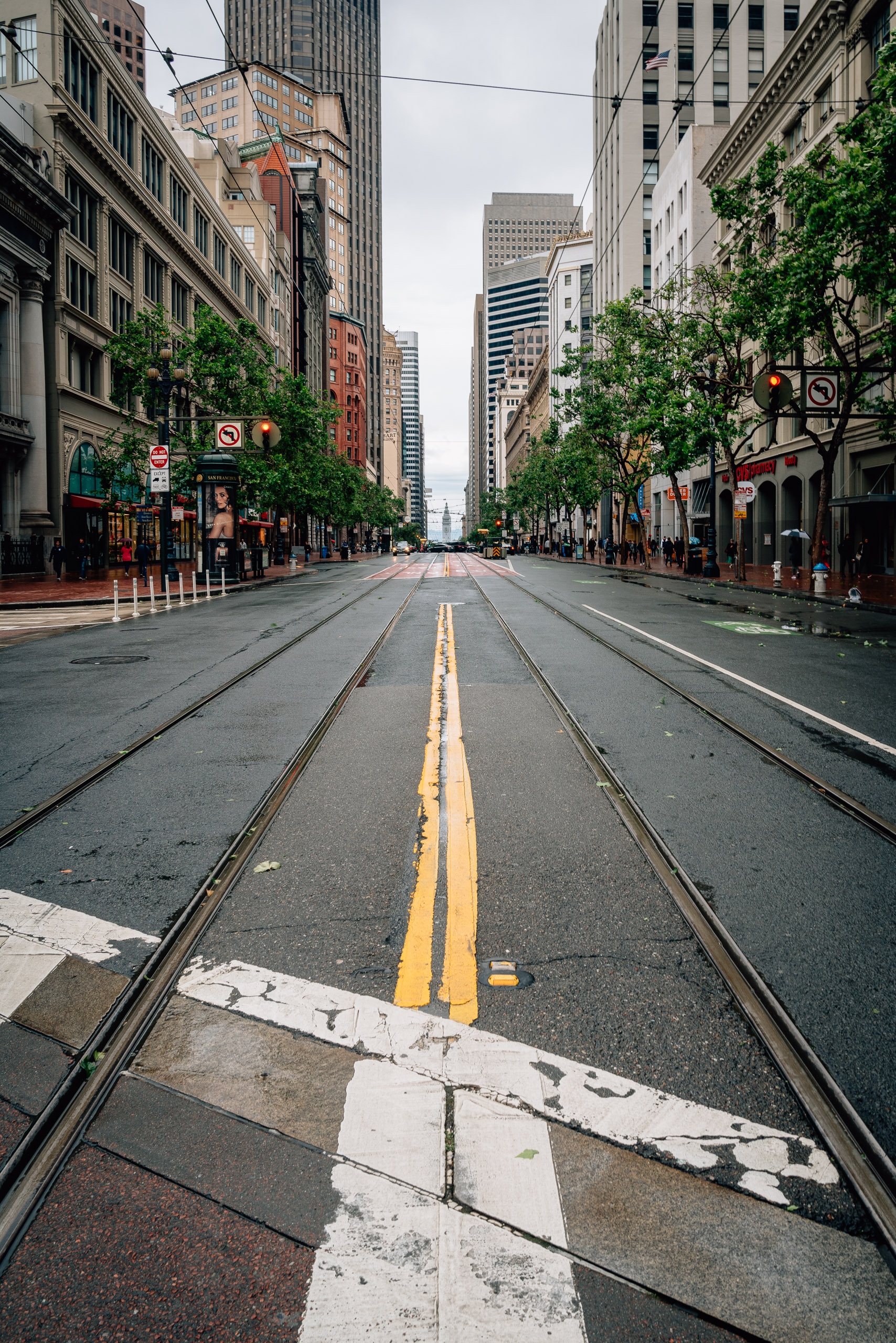 Market Street in San Francisco stretches into the distance with embedded streetcar tracks, yellow center lines, and historic buildings flanking both sides under an overcast sky
