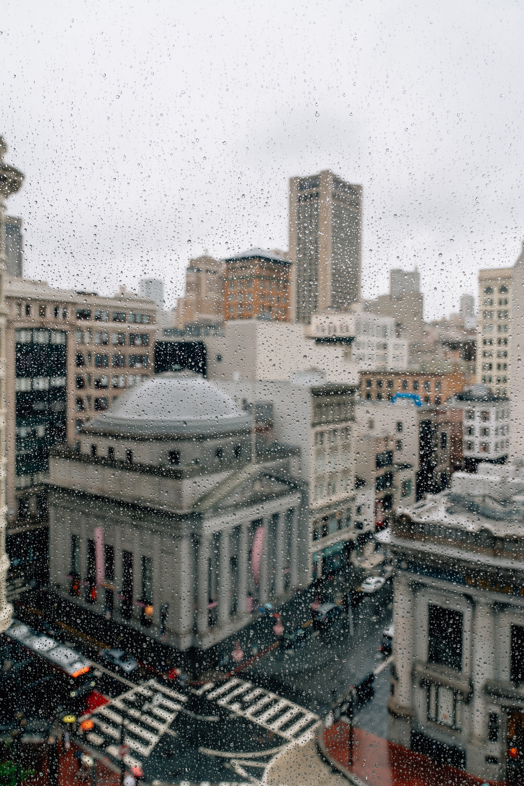 Rain-streaked window reveals a downtown San Francisco cityscape featuring a domed classical building, modern towers, and wet streets below on an overcast day