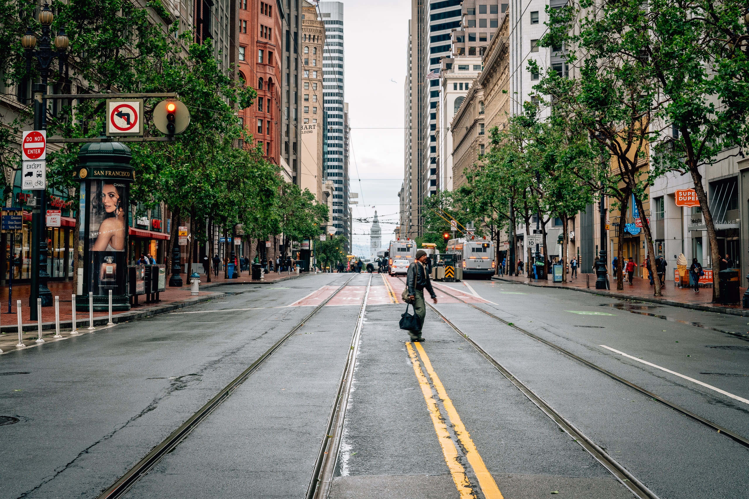 A solitary pedestrian walks down Market Street's cable car tracks lined with vine-covered buildings, shops, and towering skyscrapers under overcast San Francisco sky