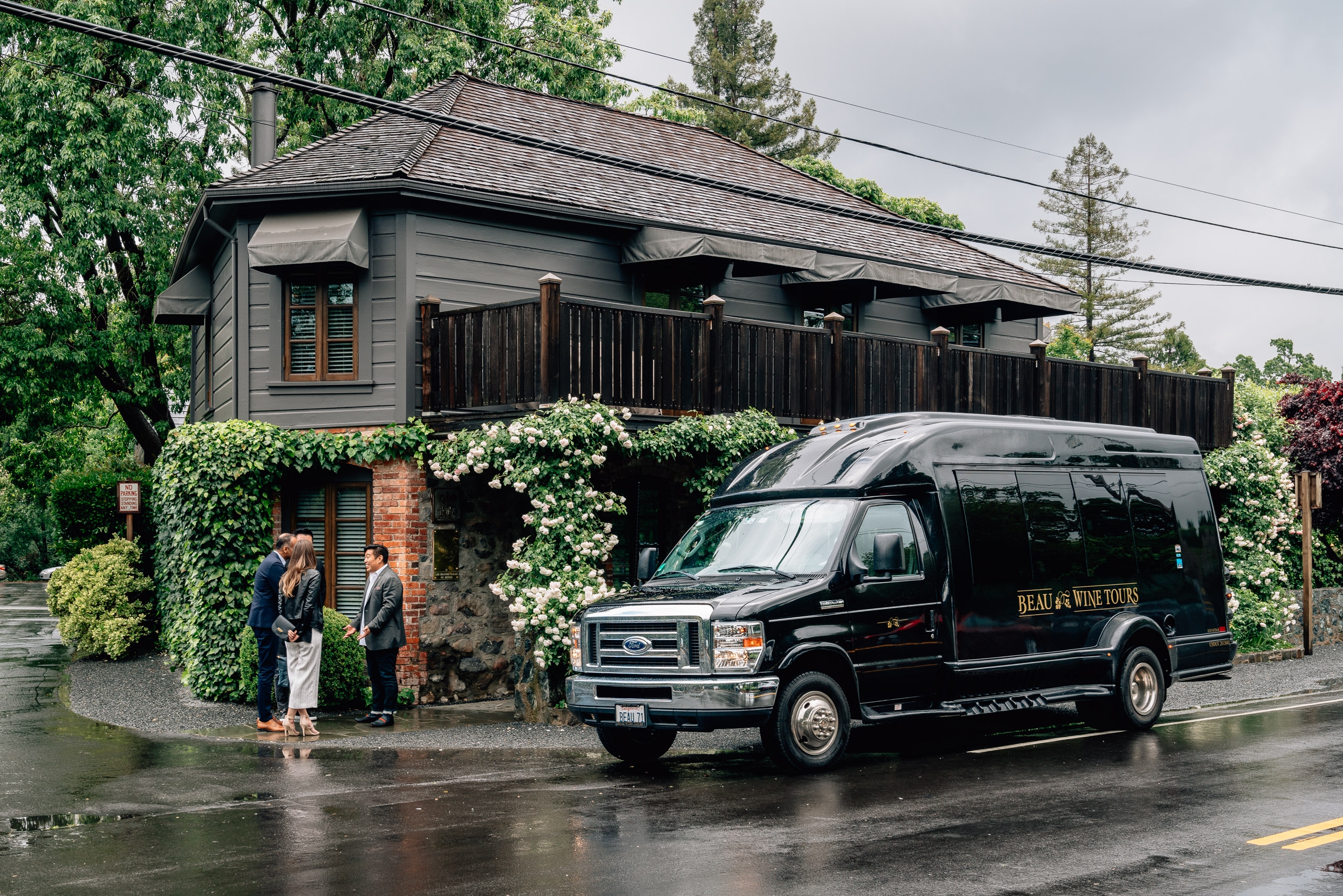 A black shuttle van labeled "Beaulieu Wine Tours" parks outside The French Laundry restaurant, with two people standing under the ivy-covered entrance during overcast, rainy conditions