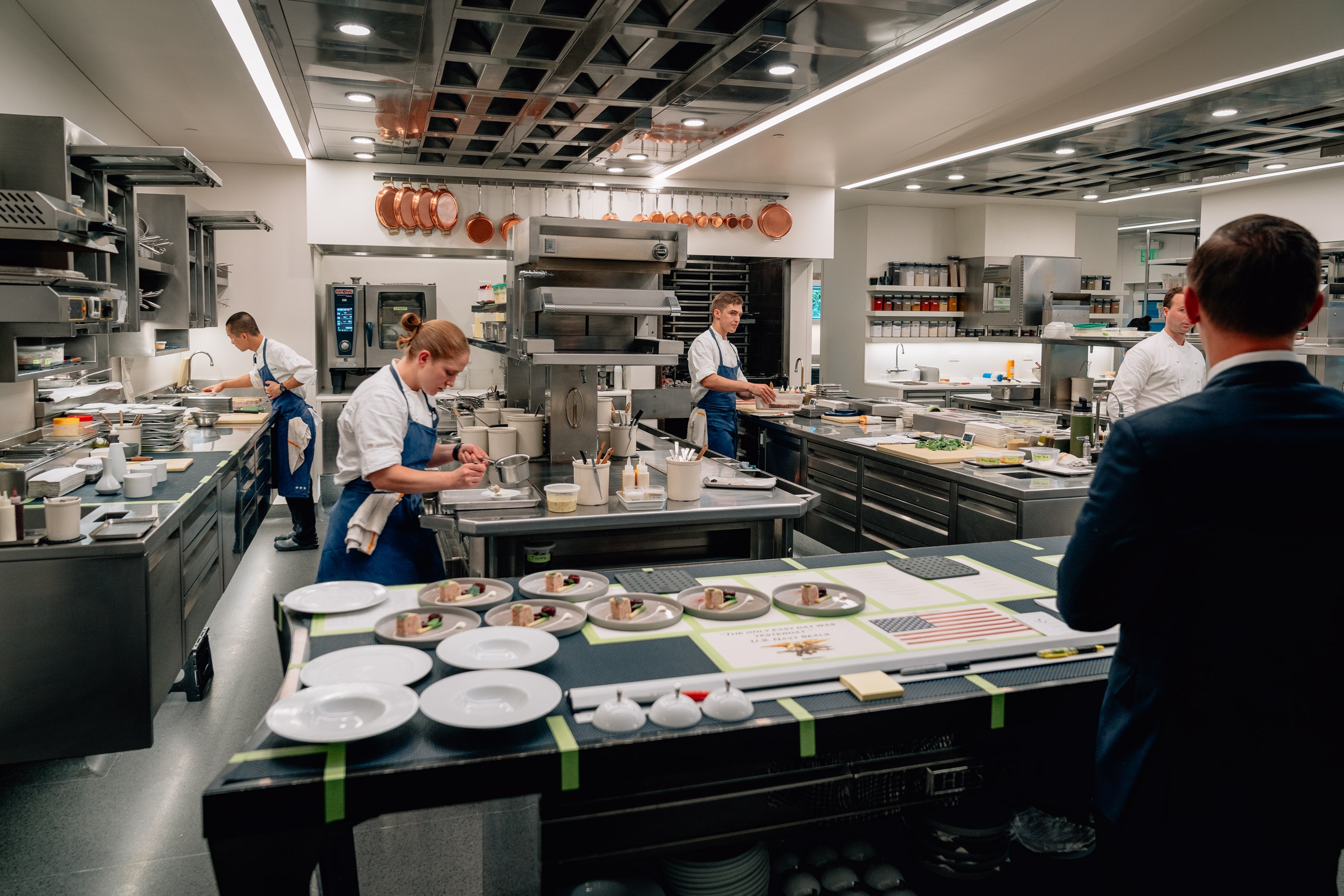 Professional chefs in blue aprons work across a bright, modern kitchen at The French Laundry, plating elegant dishes on white plates arranged on a central counter