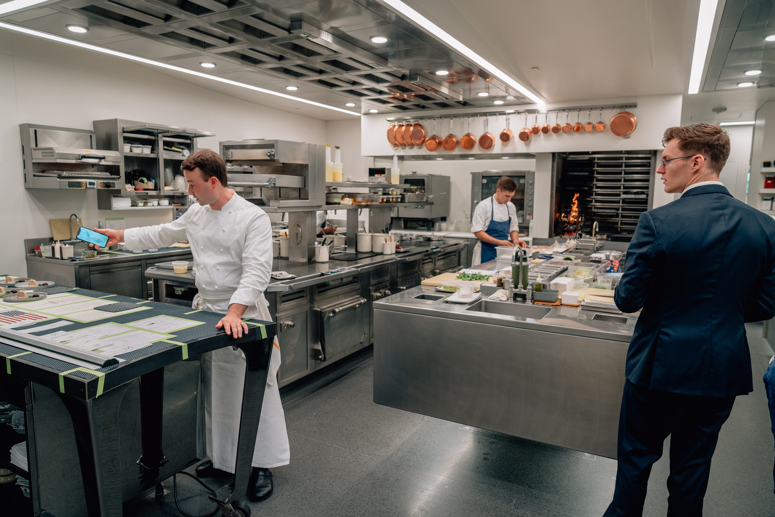 Professional chefs work in a modern kitchen with stainless steel equipment and copper cookware hanging overhead, while a suited man observes the culinary preparation