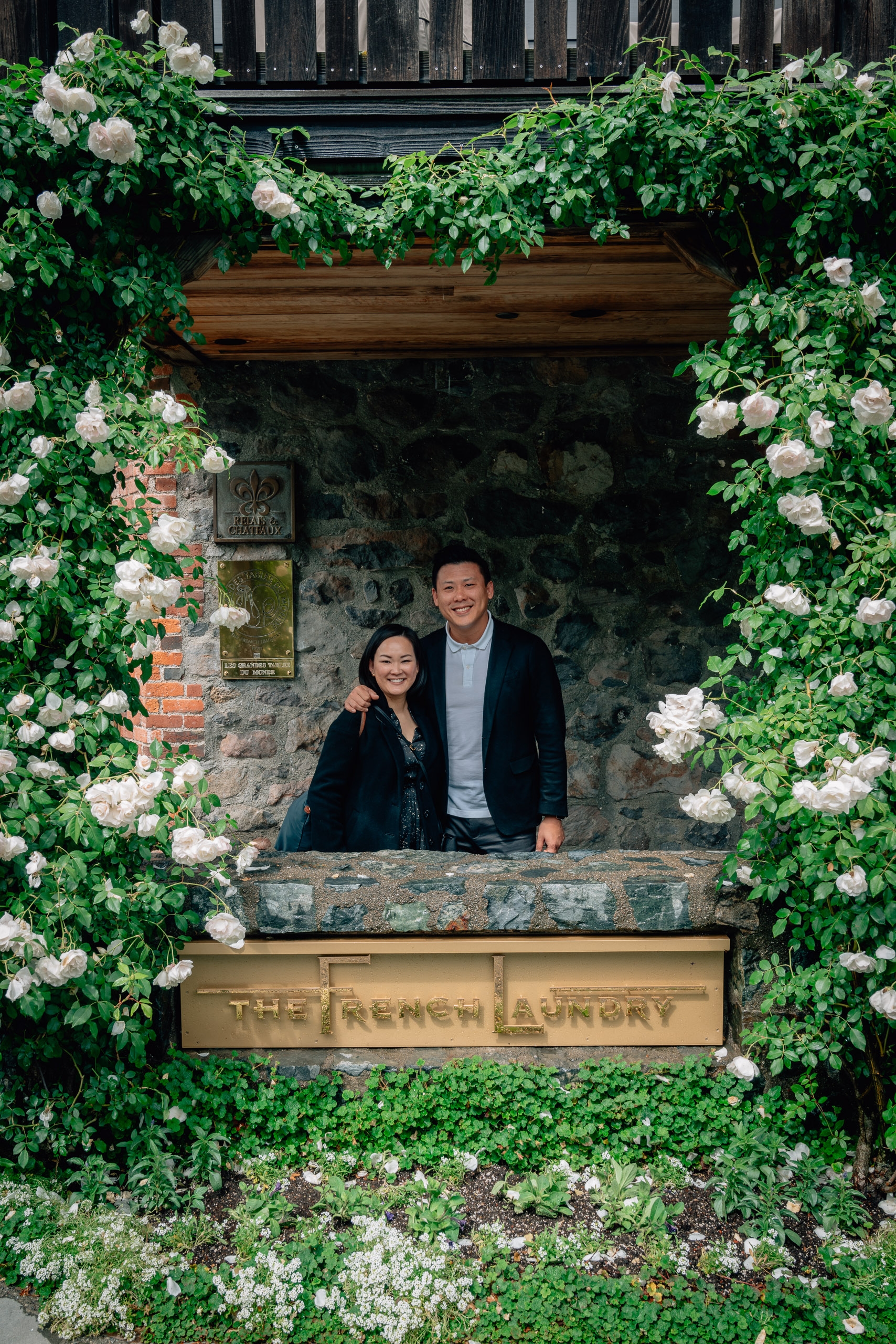 A couple stands smiling in a historic stone window frame draped with lush white flowering vines and green ivy at The French Laundry restaurant in Yountville