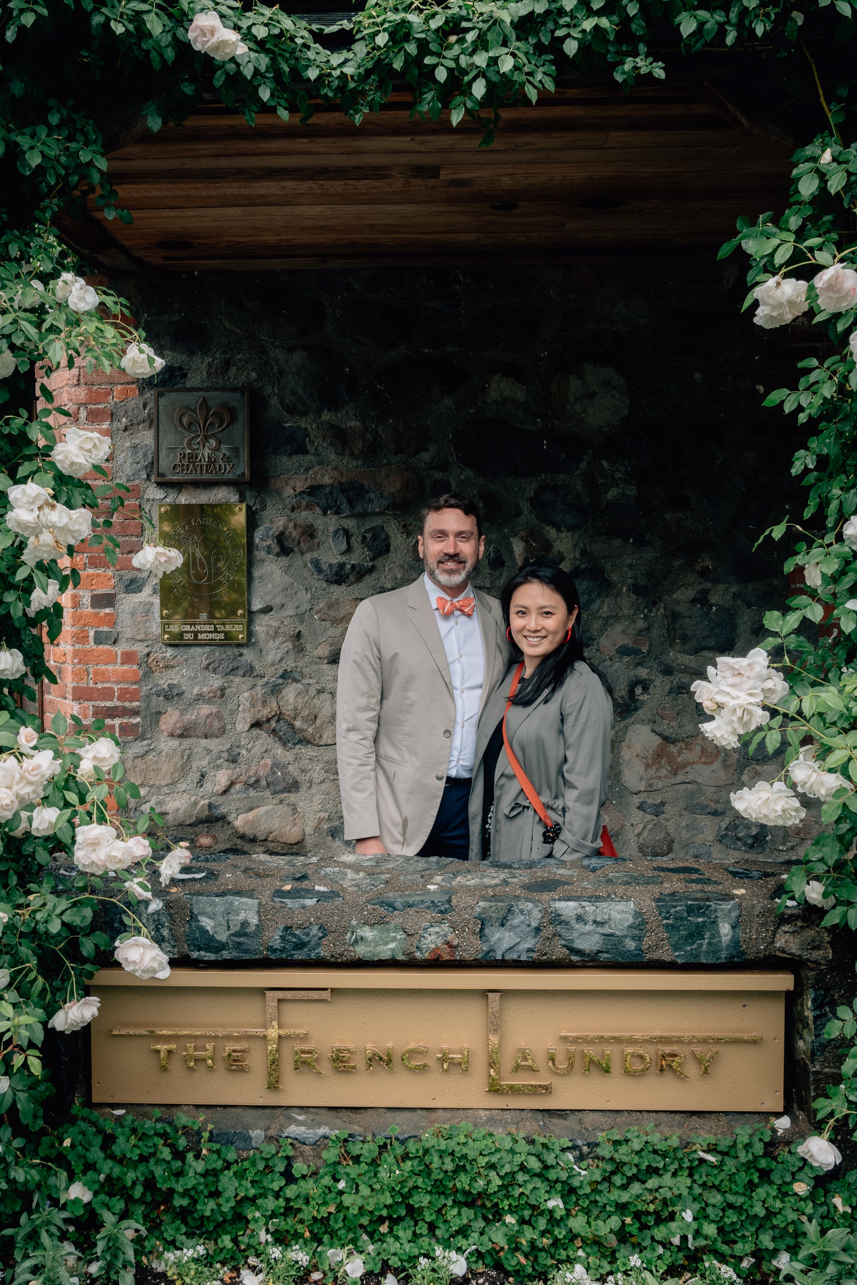 A couple stands framed within an ivy-covered stone archway at The French Laundry, surrounded by white flowering vines and weathered brick walls