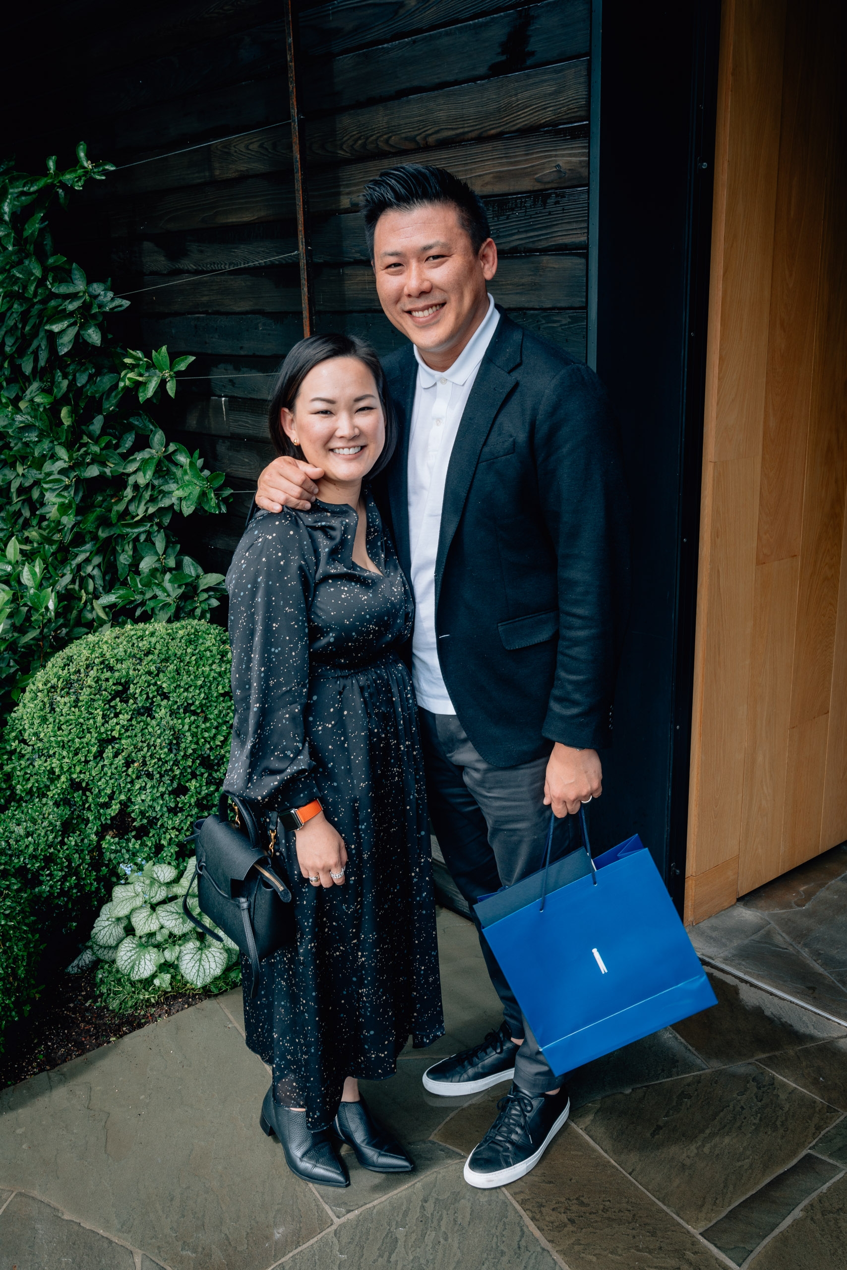 A smiling couple poses together outside a modern doorway, with the man holding a blue shopping bag and the woman wearing a dark sparkly dress, framed by lush green ivy landscaping