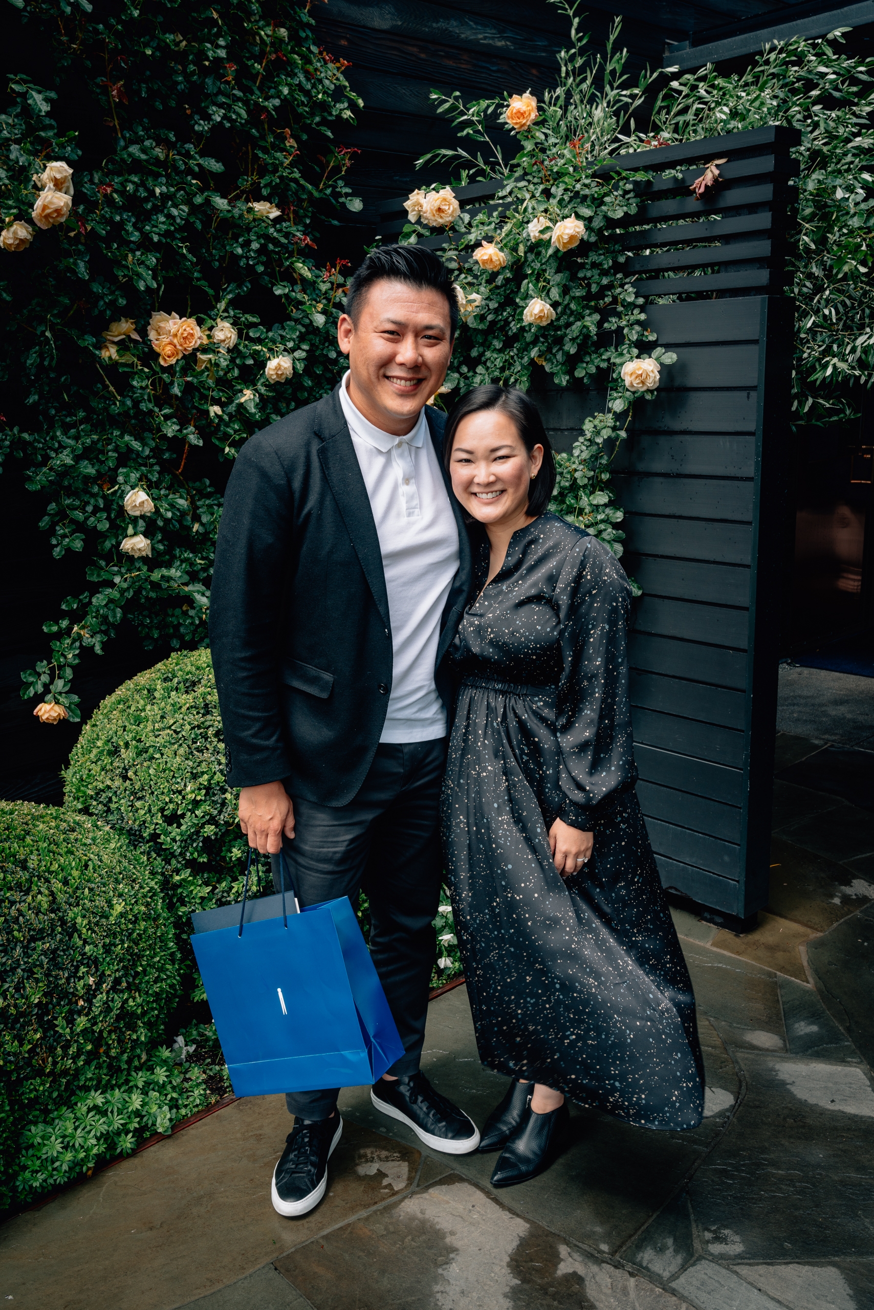 A smiling couple poses together in front of a lush green floral wall adorned with peachy-orange roses, the man holding a blue shopping bag in formal attire