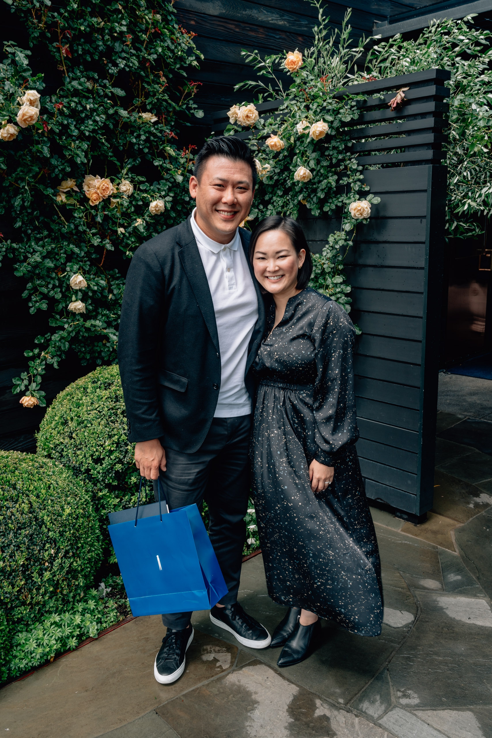 A smiling couple stands together against a lush green floral backdrop adorned with peach-colored roses, with the man holding a bright blue shopping bag