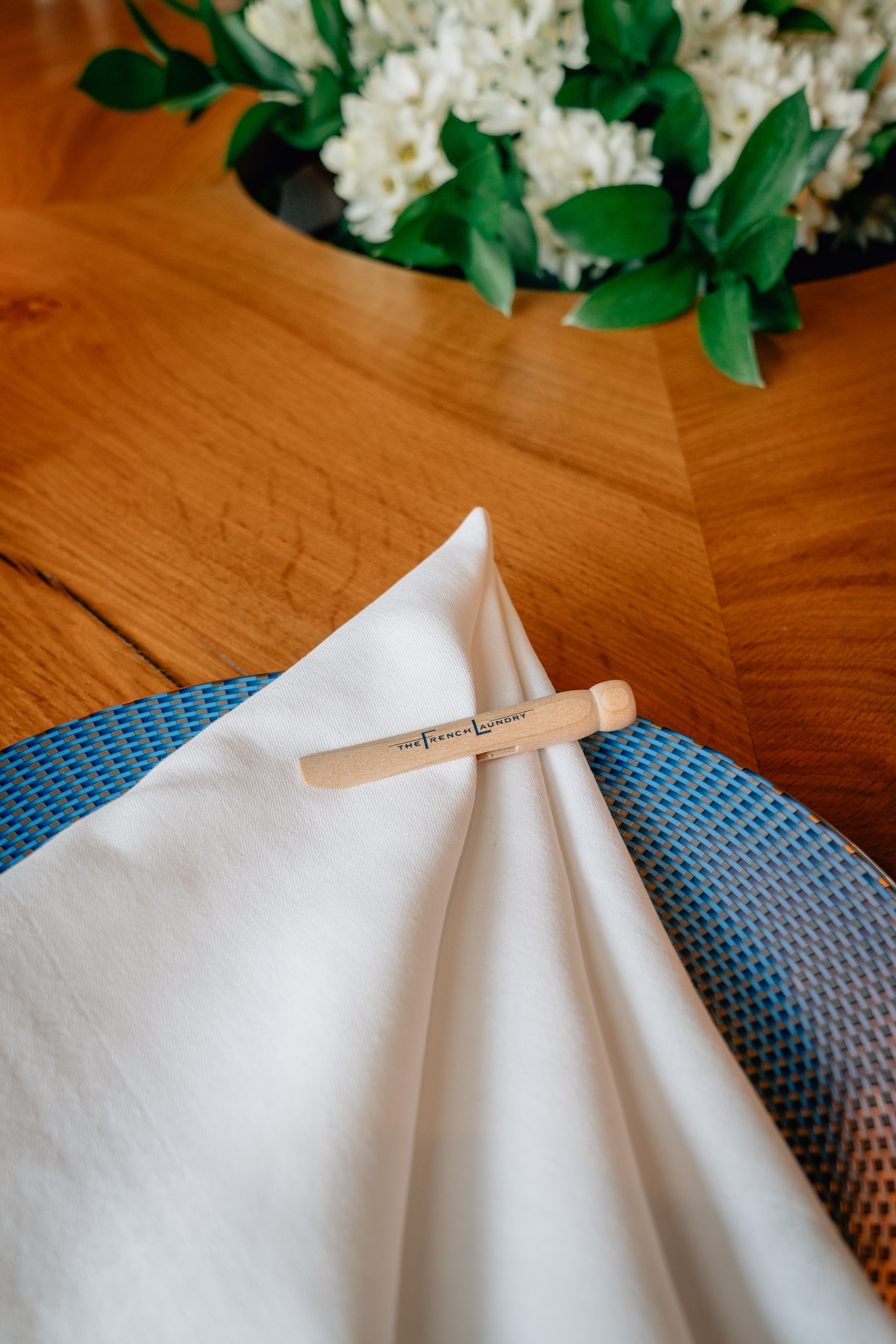 A wooden table displays a formal place setting with a white folded napkin, wooden utensil, and blue textured placemat, with white flowers and greenery in a vase above