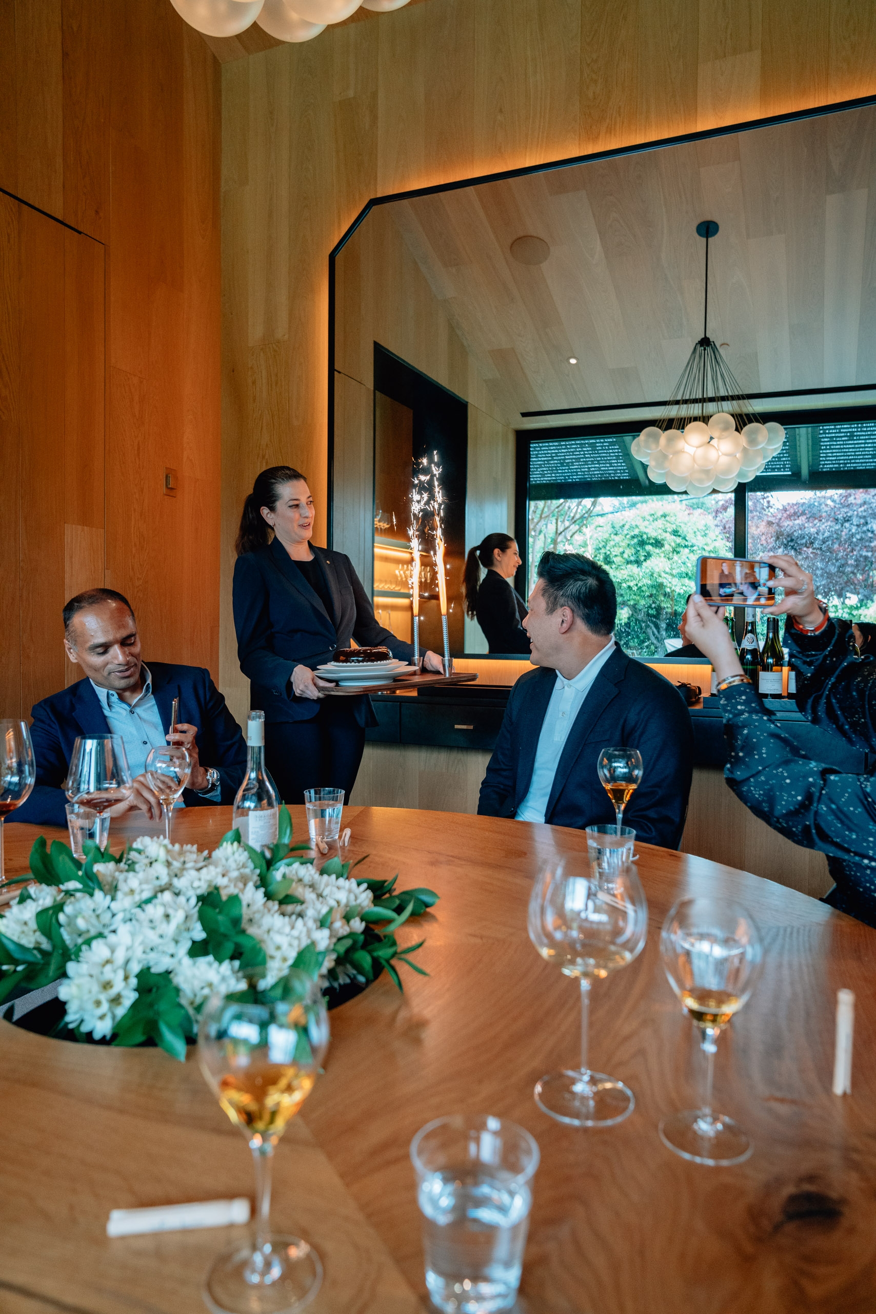 A woman serves drinks at a formal dining table where suited men gather, with white flowers and glassware visible under warm wood paneling and a cloud-like ceiling fixture
