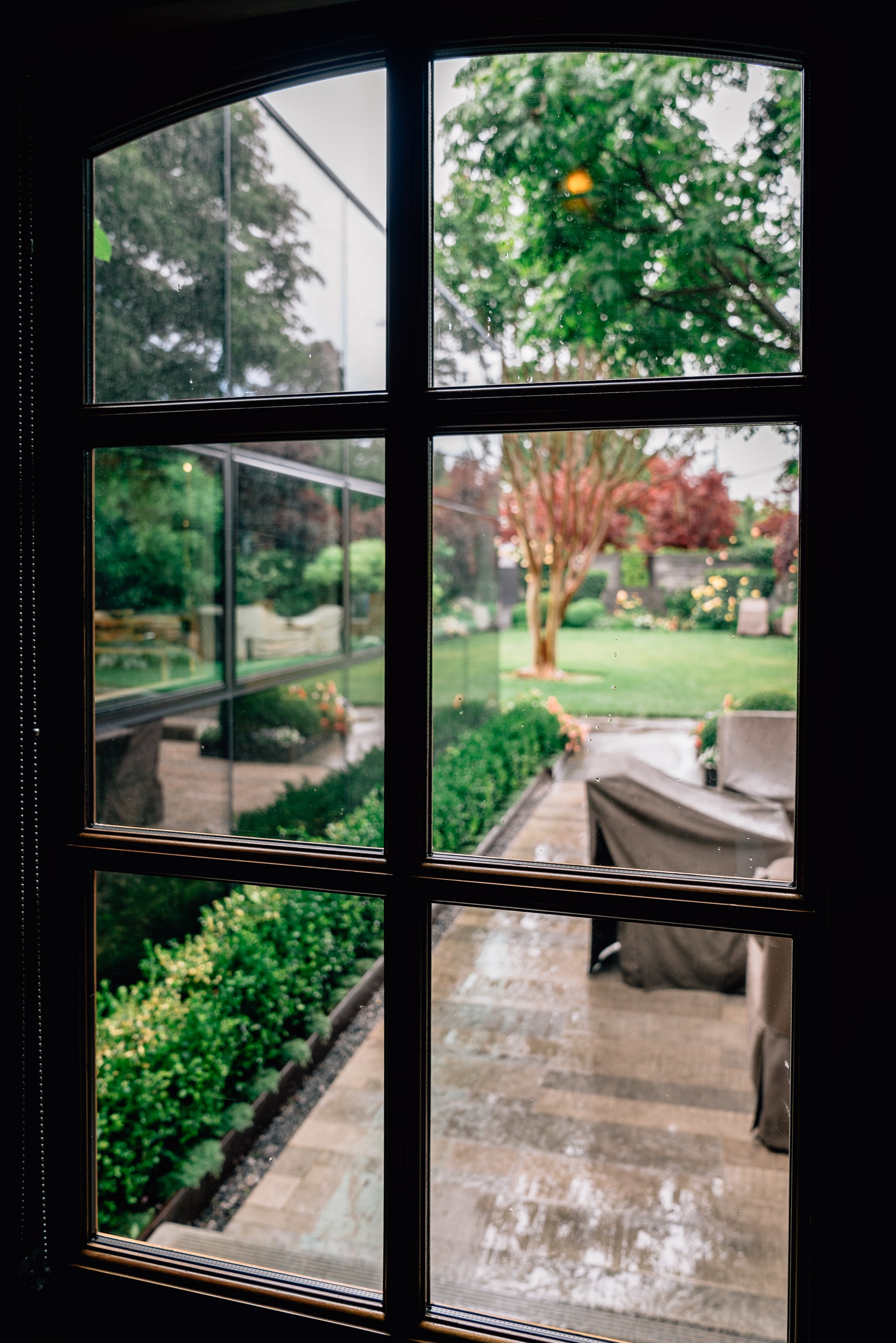 Six-pane window framing views of a manicured garden with green hedges, lawn, and a flowering tree in soft afternoon light