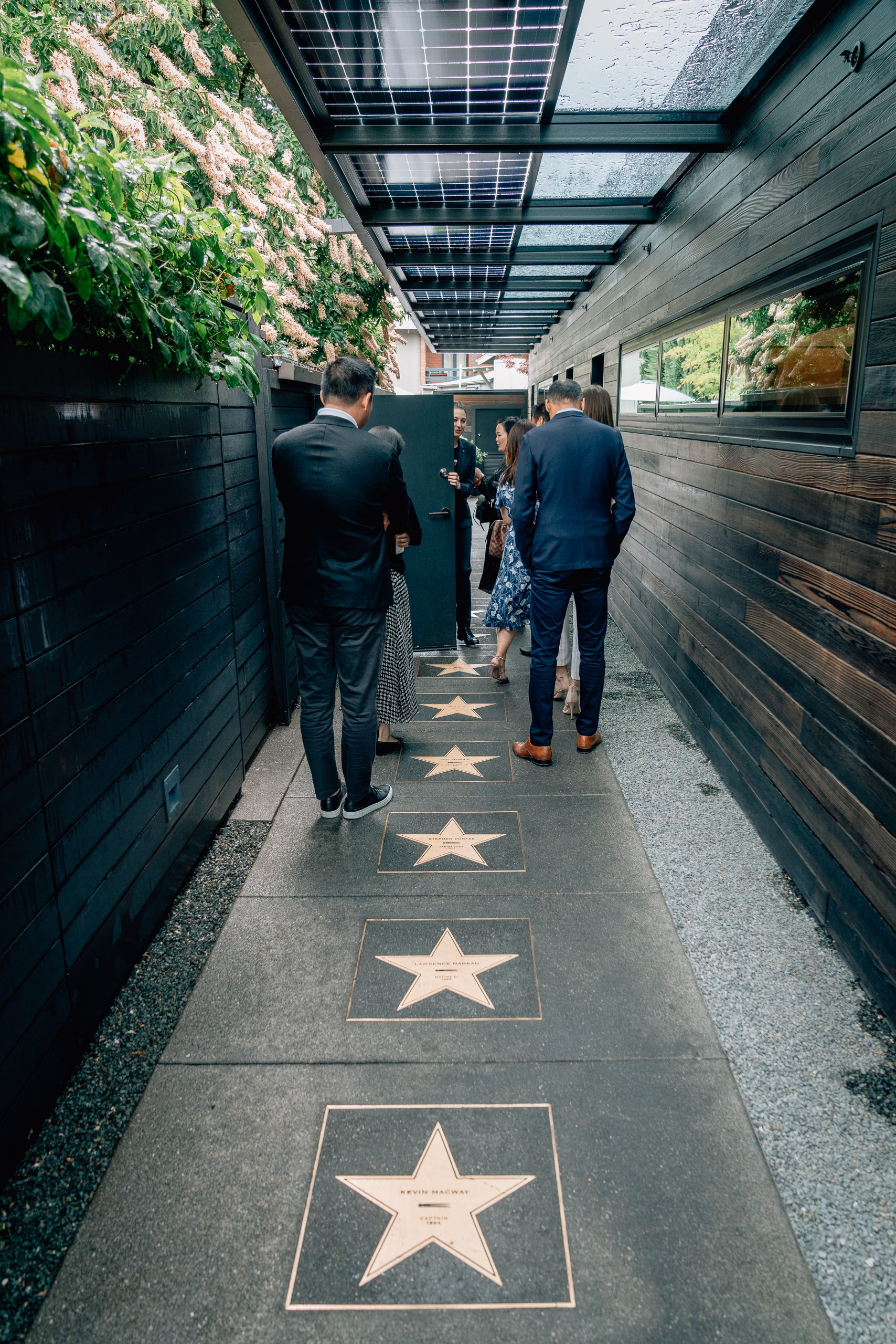 Elegantly dressed visitors walk along the Hollywood Walk of Fame's star-lined pavement beneath solar panel skylights and ivy-covered walls in a narrow corridor