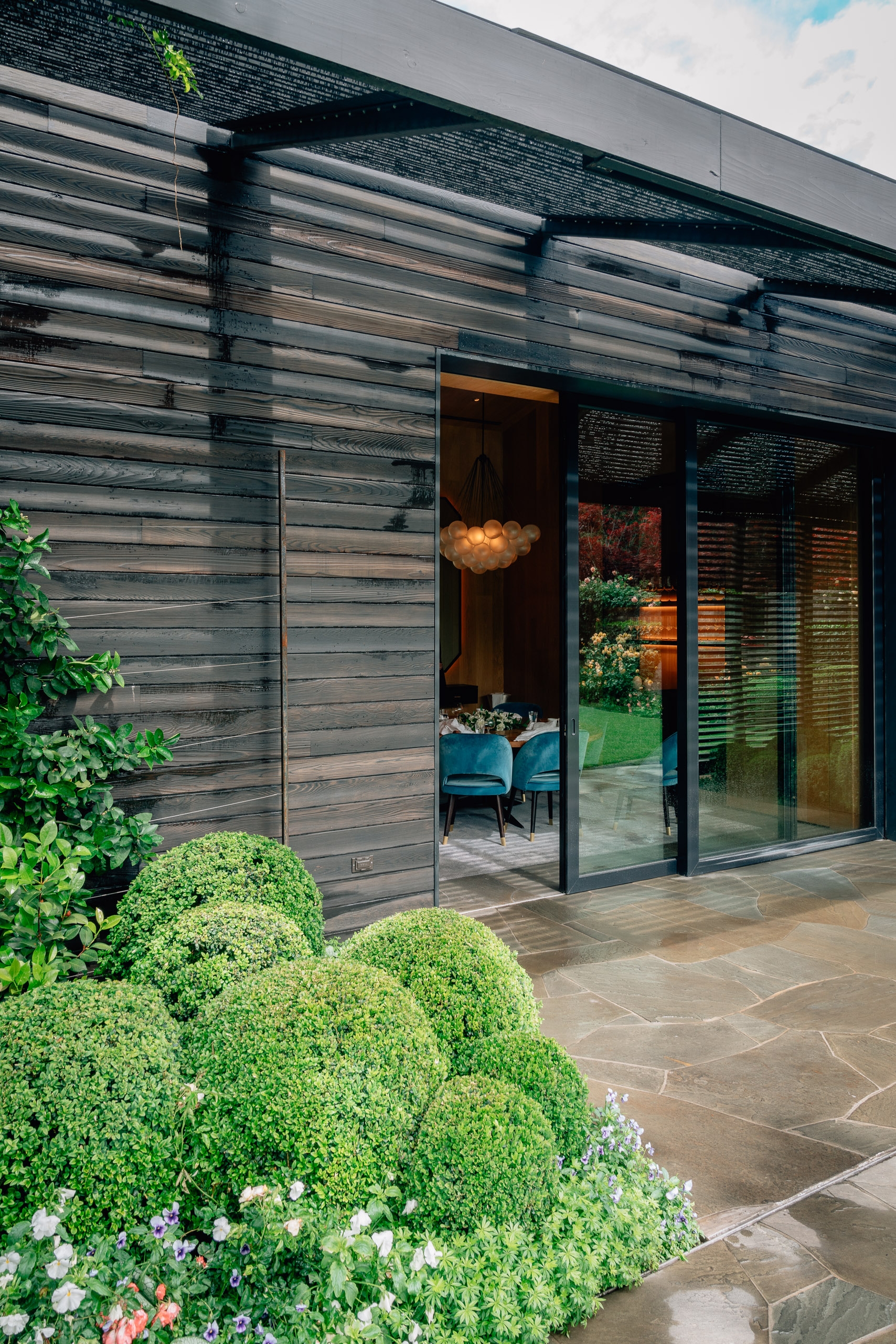 Modern dark wood-clad structure with glass sliding doors opens onto a patio, featuring manicured green hedges and flowering plants in the foreground under bright daylight