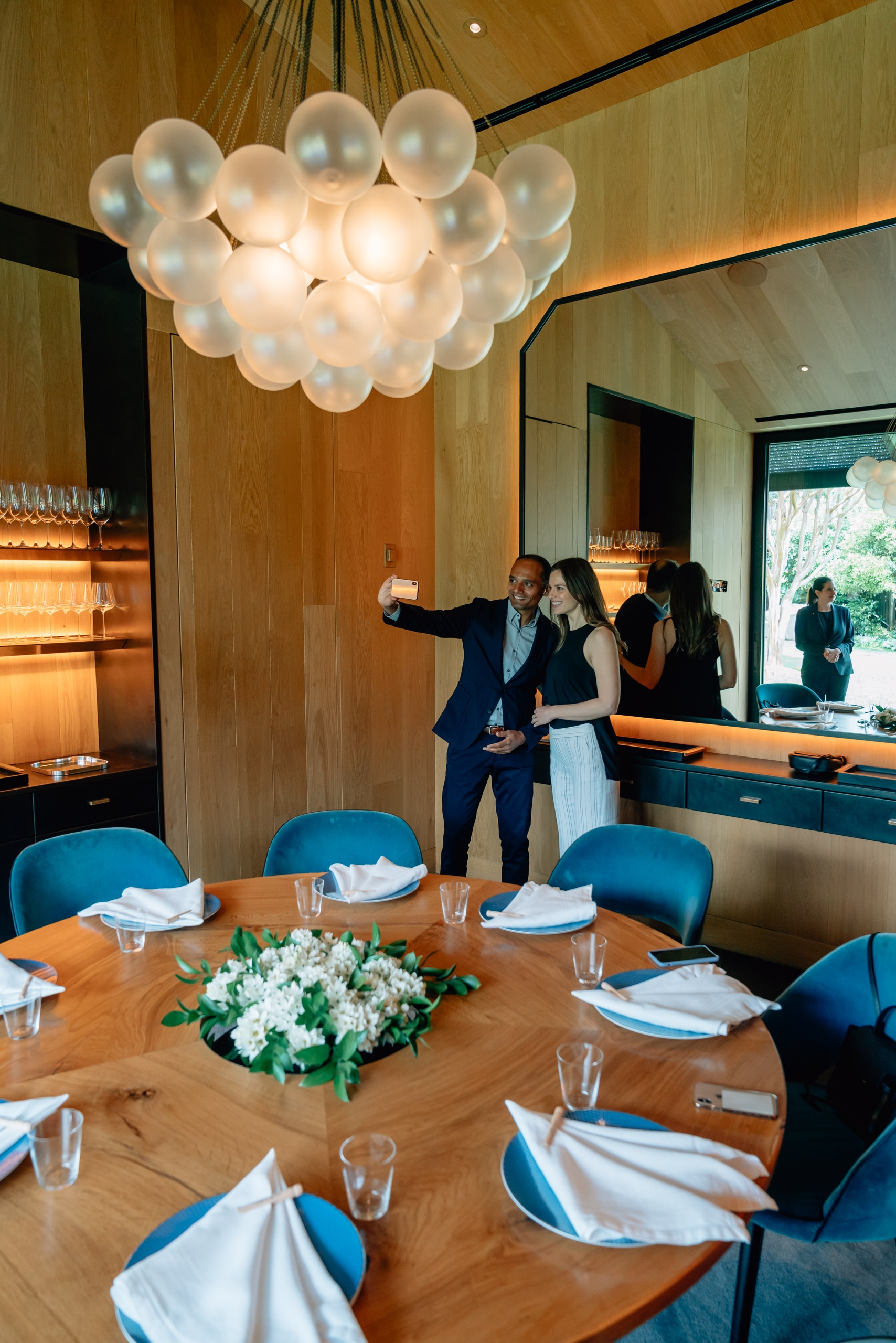 A modern dining room features a large spherical pendant light above a wooden table set with place settings and white flowers, while two people stand near the bar area in the background.