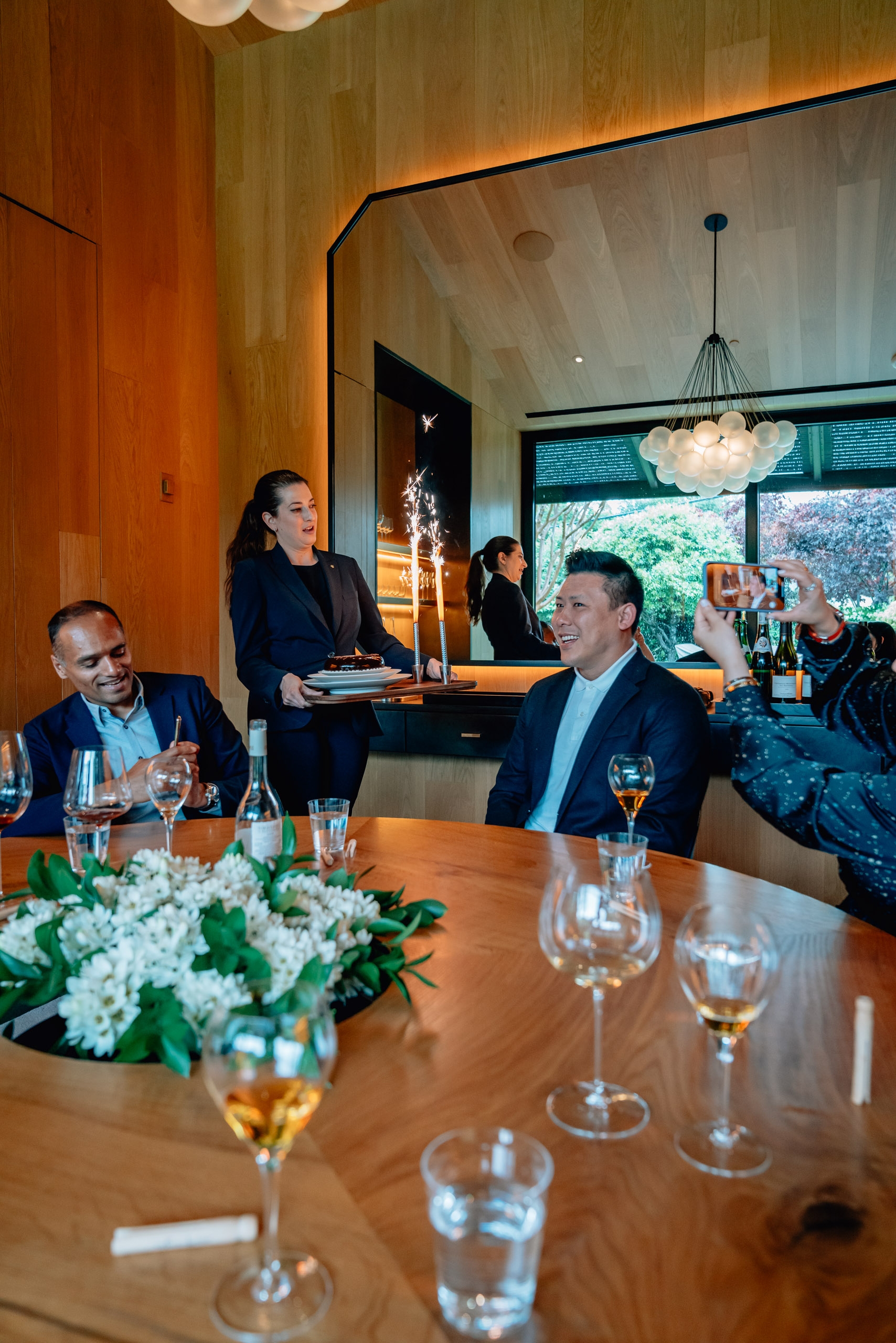 Professional diners gather around a wooden table in a modern restaurant, with wine glasses and white flowers in the foreground, warm wood paneling, and contemporary pendant lighting overhead
