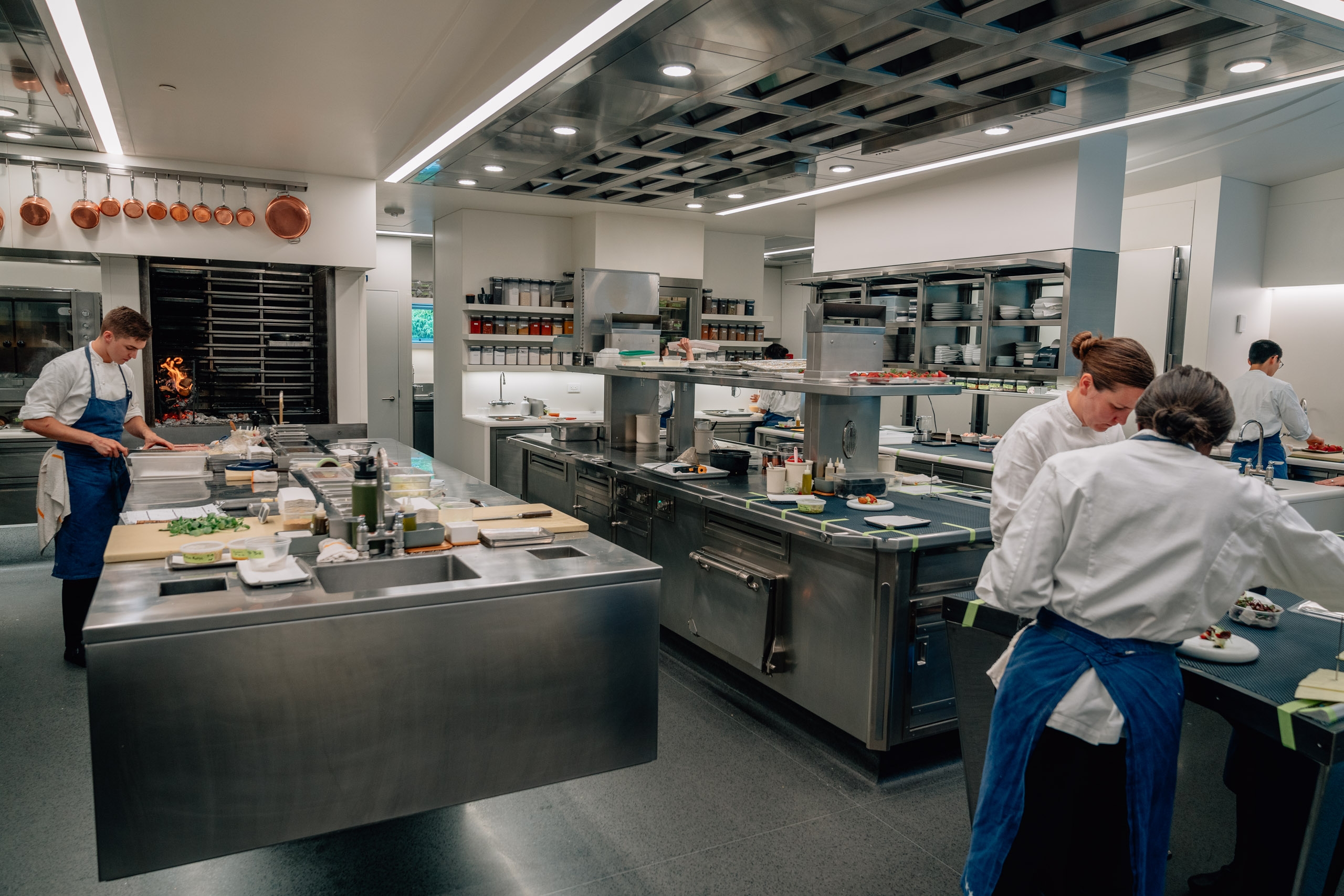 Professional chefs work at stainless steel stations in a bright, modern restaurant kitchen with organized shelving, hanging copper cookware, and recessed lighting overhead