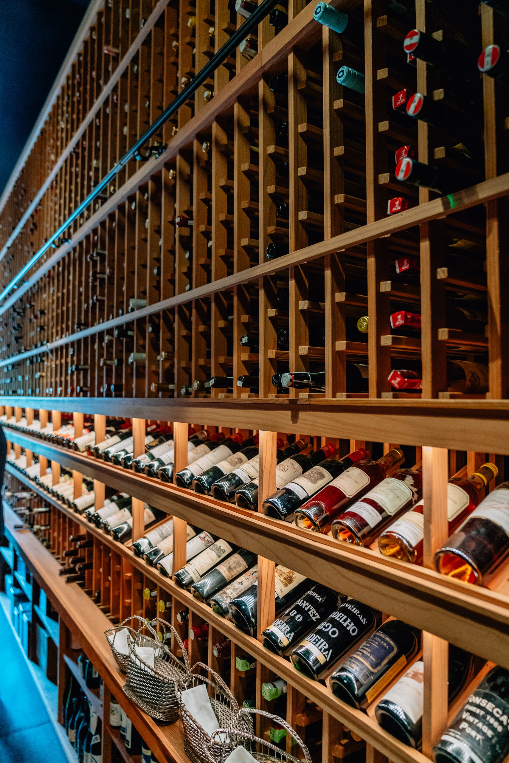 A modern wine cellar displays hundreds of bottles organized in wooden racks with blue accent lighting, showcasing white and red wines stored horizontally across multiple tiers