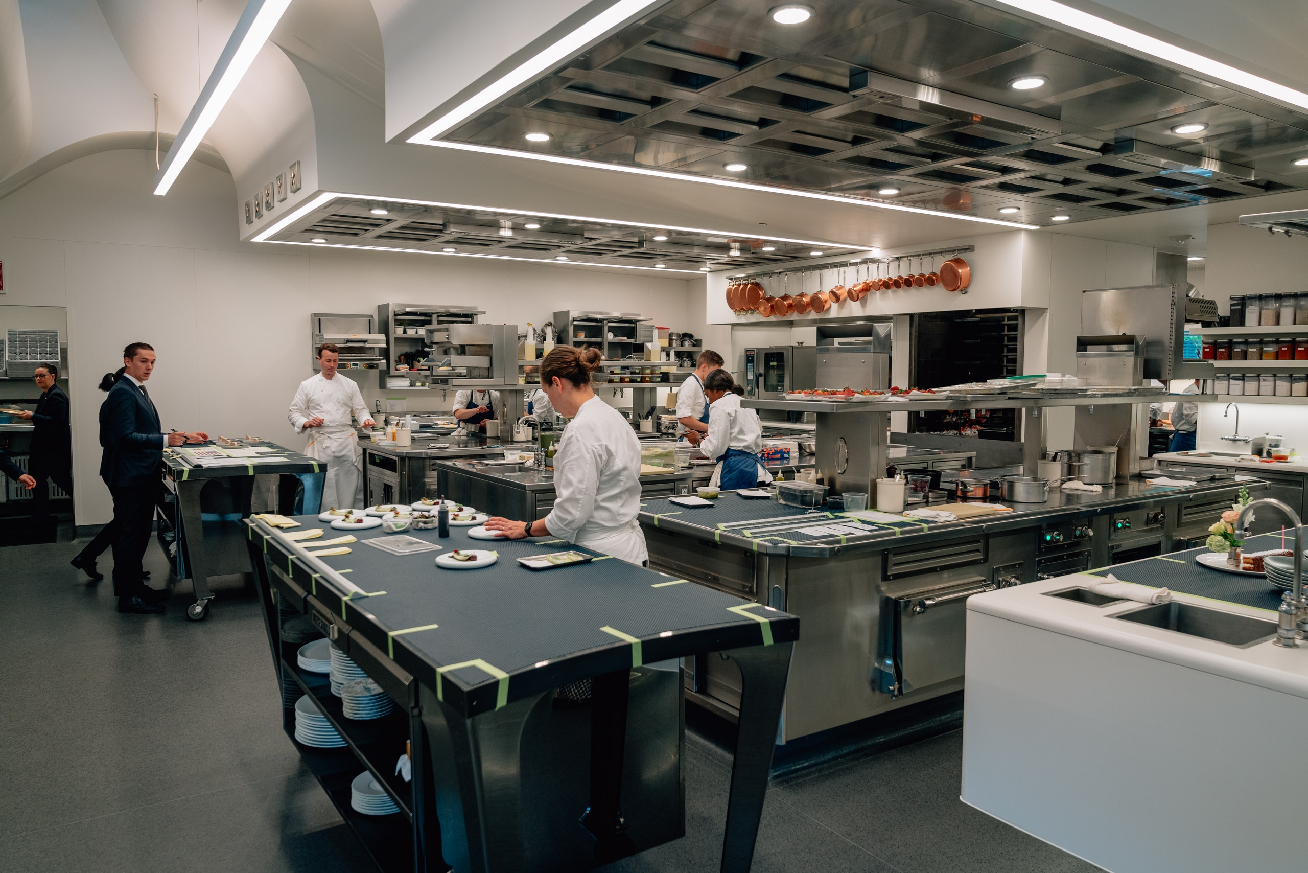 Professional chefs work at stainless steel stations in a modern, brightly lit commercial kitchen with linear ceiling fixtures and industrial equipment