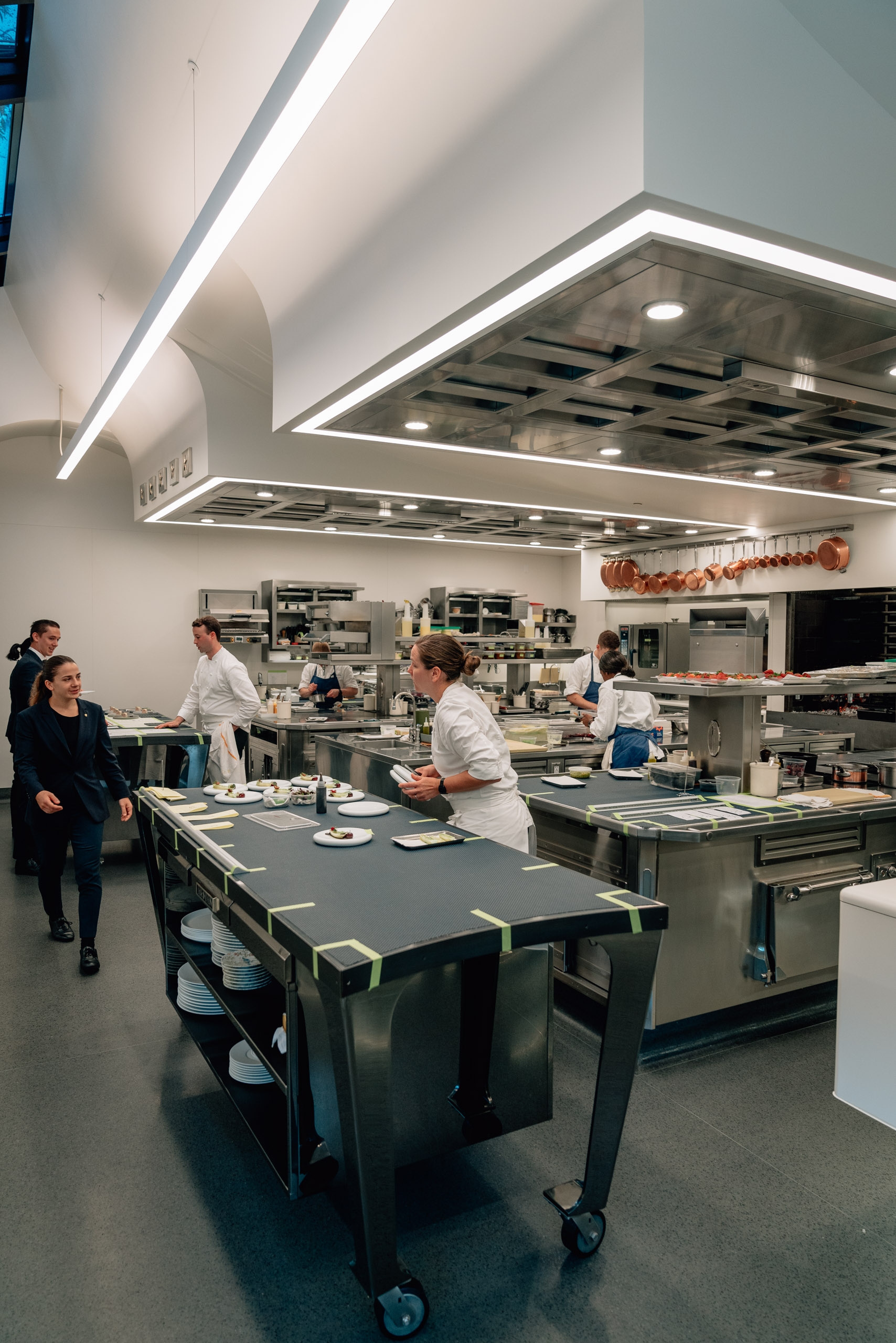 Professional chefs and kitchen staff work at multiple stations in a bright, modern culinary kitchen with stainless steel equipment and linear ceiling lights overhead