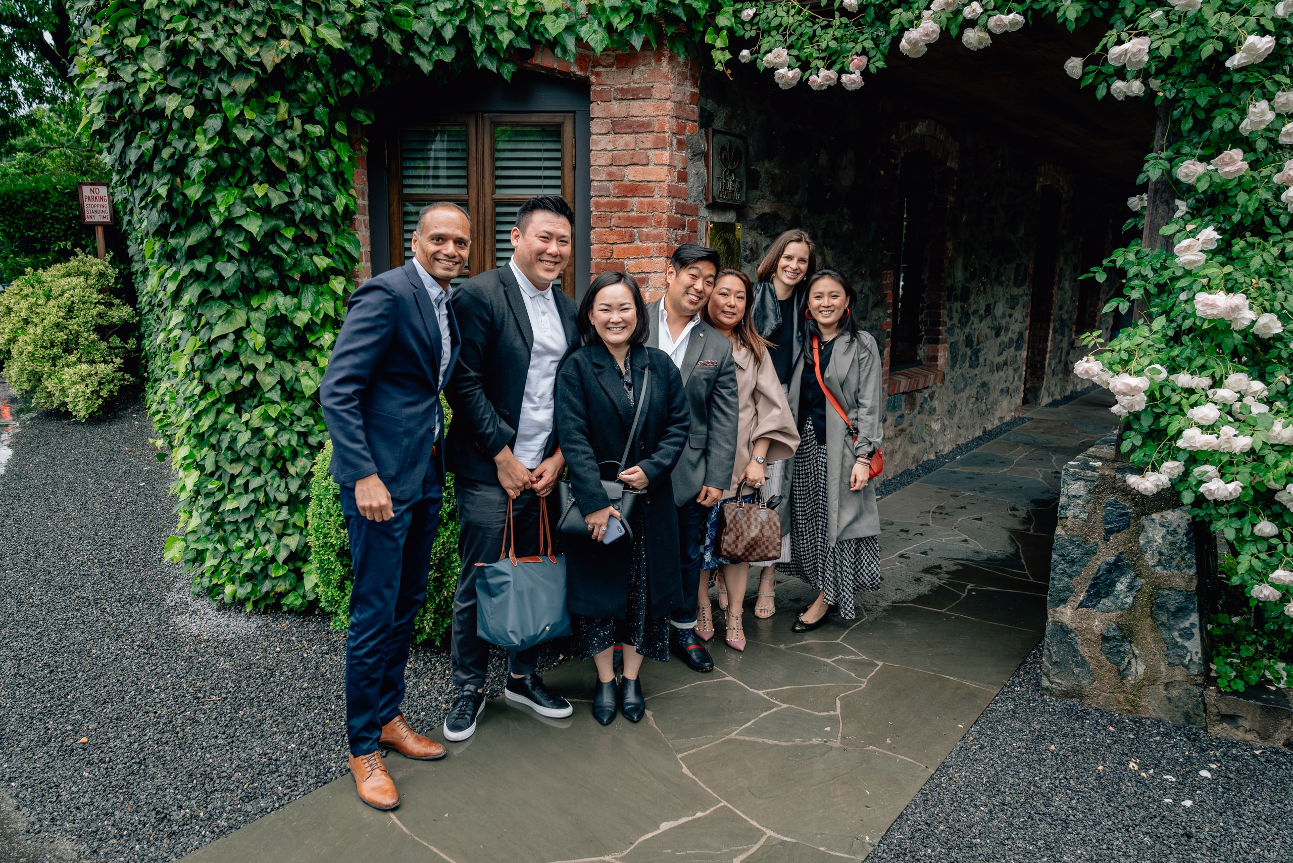 A group of eight professionally dressed people stands together in a charming ivy-covered passageway with white flowering vines and brick architecture, smiling at the camera in daylight