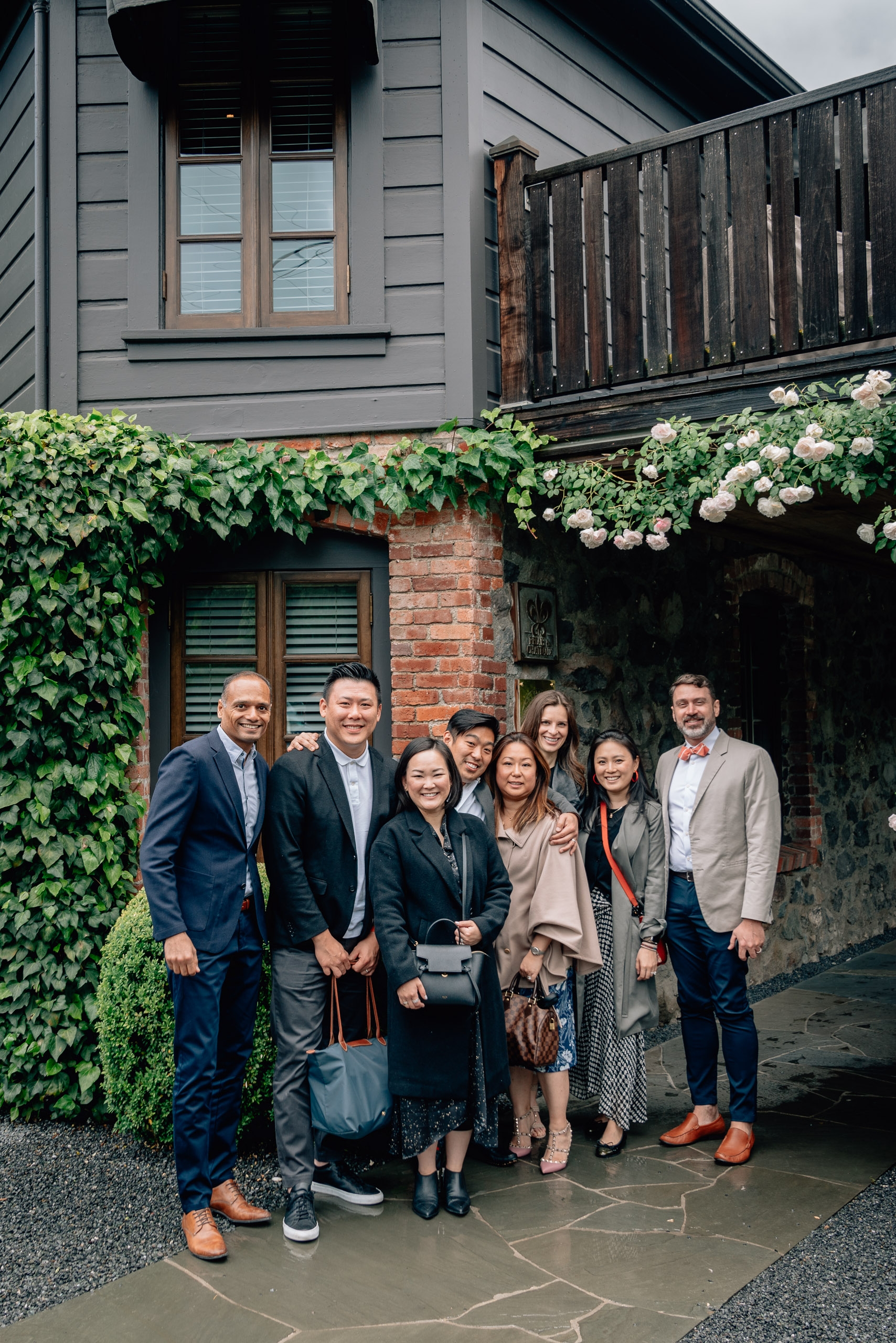 A group of nine professionally dressed people stand together in front of an ivy-covered brick archway with white flowering vines, posing for a formal portrait on a stone patio