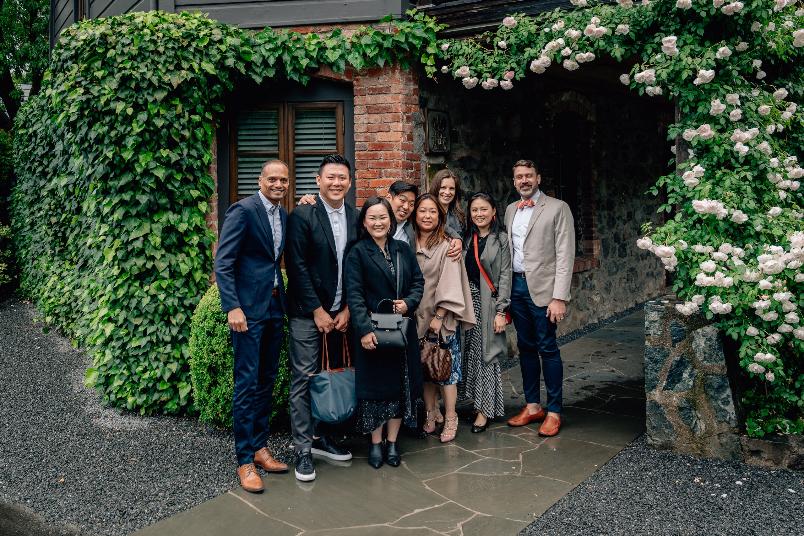A group of nine professionals dressed in business casual attire stand together under a vine-covered brick archway adorned with white flowering climbing plants