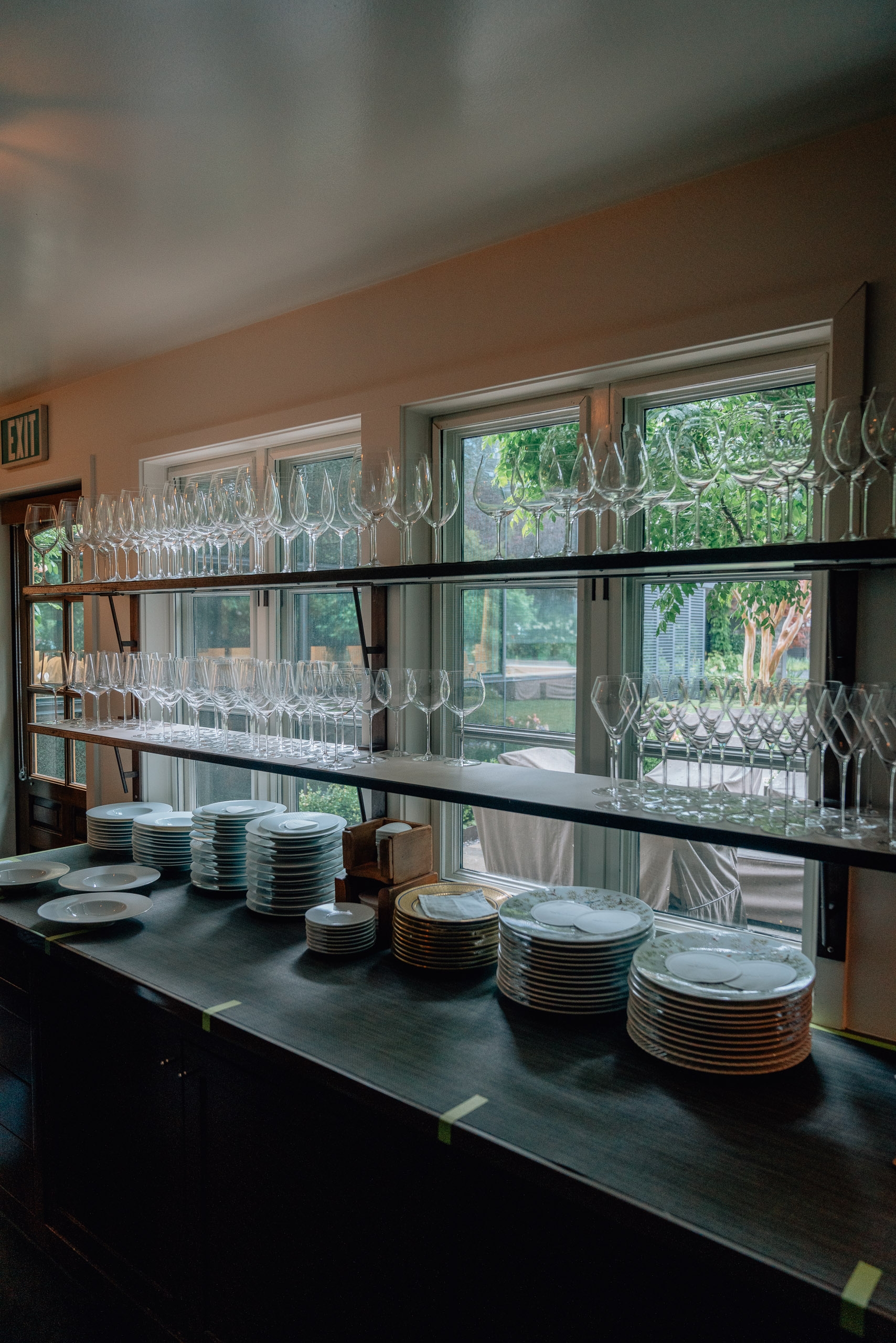 A restaurant service station displays stacked white plates and wine glasses on dark shelving beside large windows overlooking lush greenery, with peach walls and natural daylight flooding the organized kitchen workspace