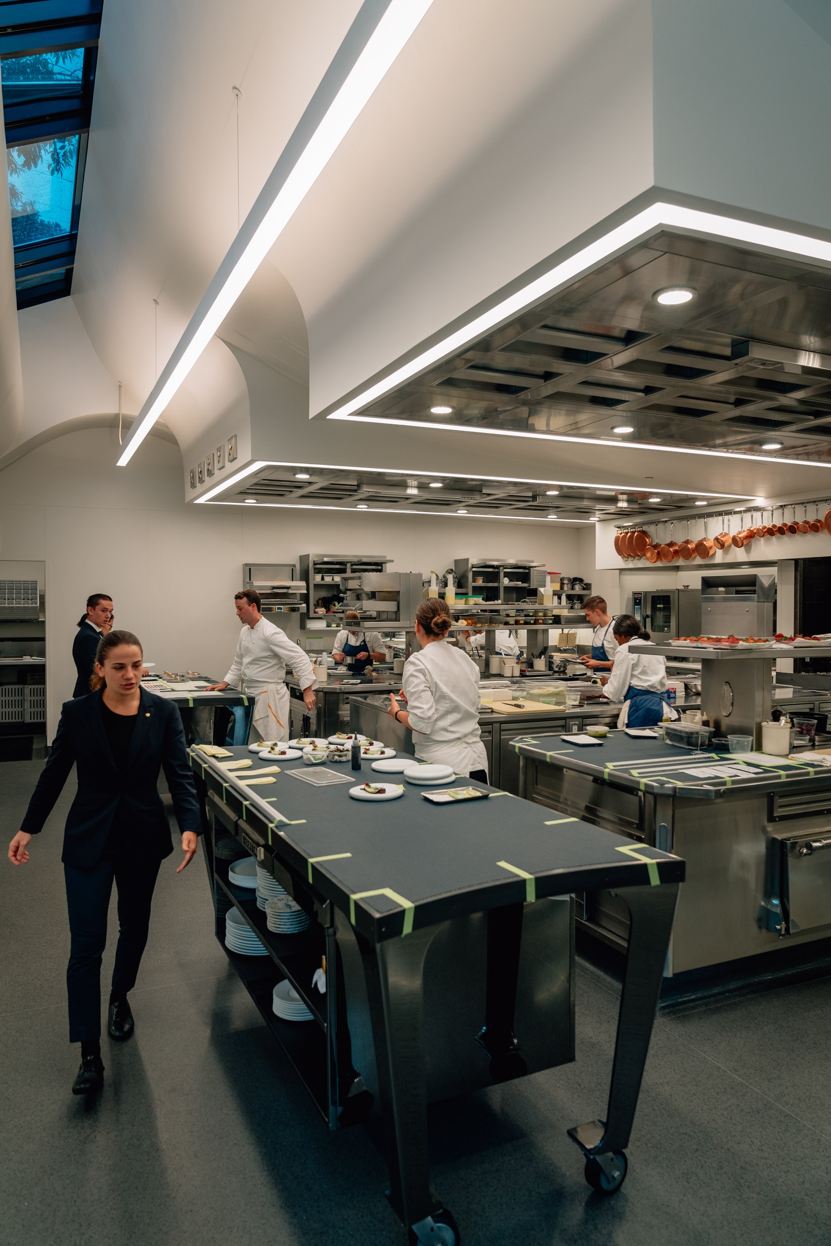Professional kitchen at The French Laundry with chefs working at central prep stations under bright linear ceiling lights and modern ventilation hoods