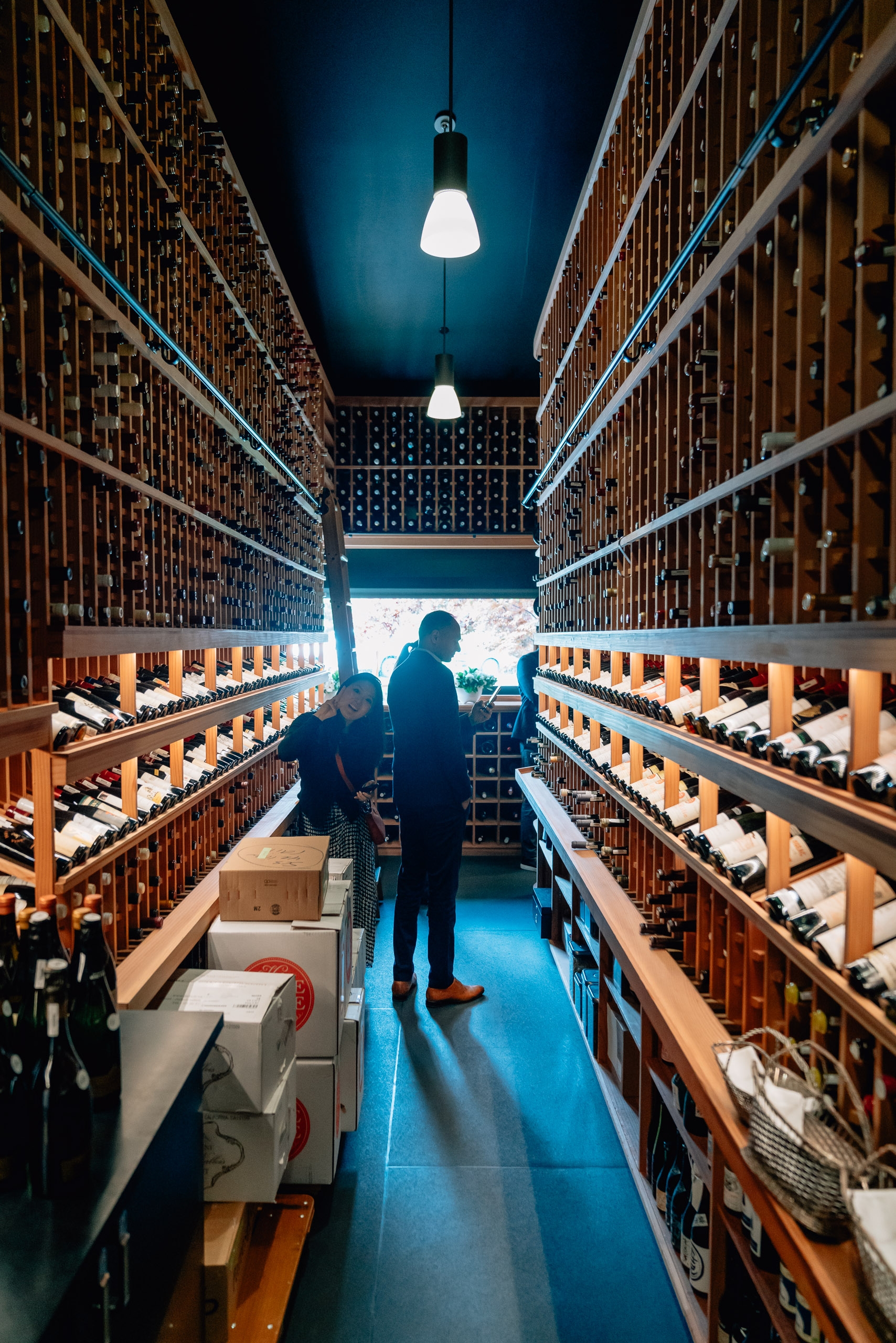 A visitor stands in a narrow wine cellar lined with wooden racks of bottles on both sides, illuminated by pendant lights overhead and daylight visible at the far end
