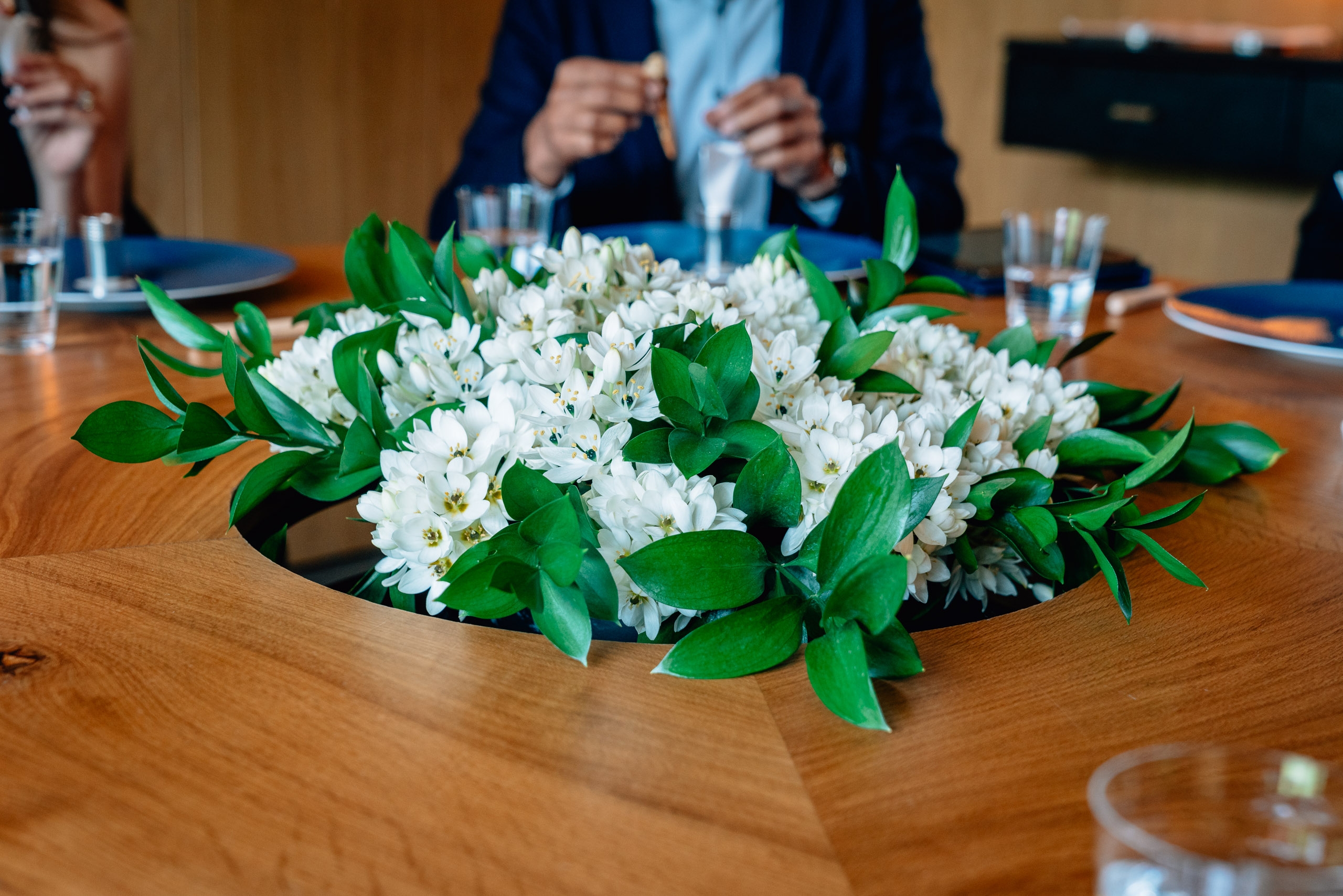 A lush arrangement of white flowers with green foliage sits as a centerpiece on a wooden conference table, with professionally dressed attendees and place settings visible in the background.