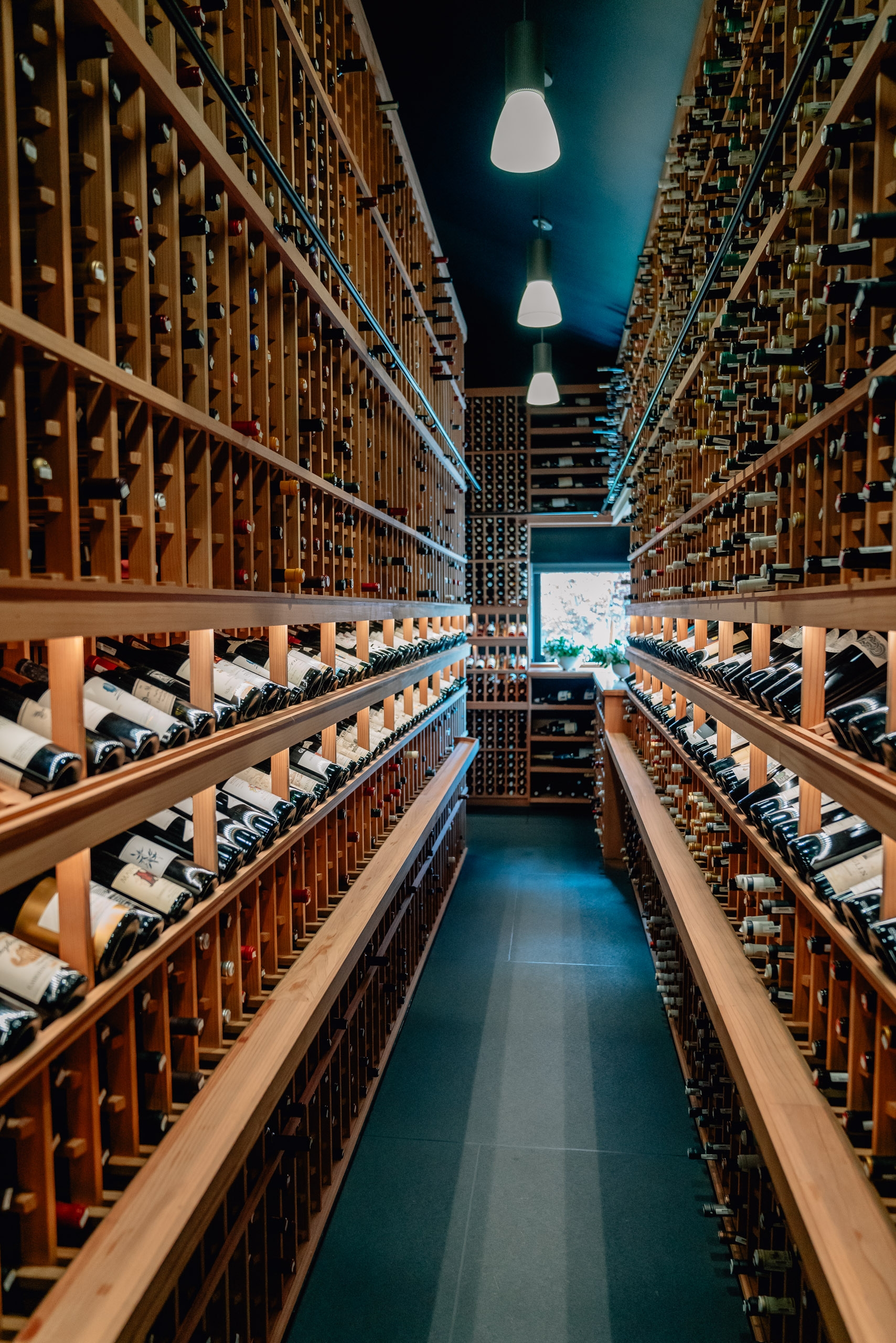 A narrow wine cellar corridor lined with wooden racks holds countless wine bottles on both sides, with pendant lights glowing overhead and a teal-blue floor stretching into the distance
