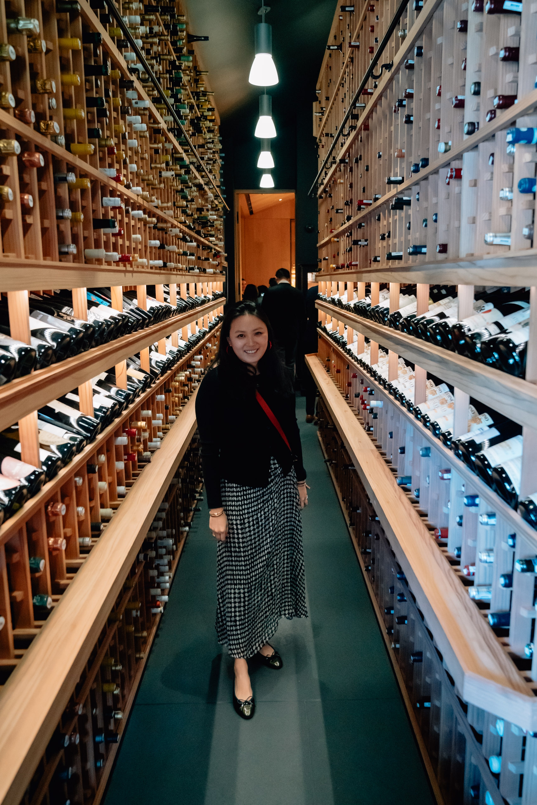 A woman stands smiling in a narrow wine cellar aisle lined with hundreds of bottles on wooden shelves, illuminated by pendant lights overhead.