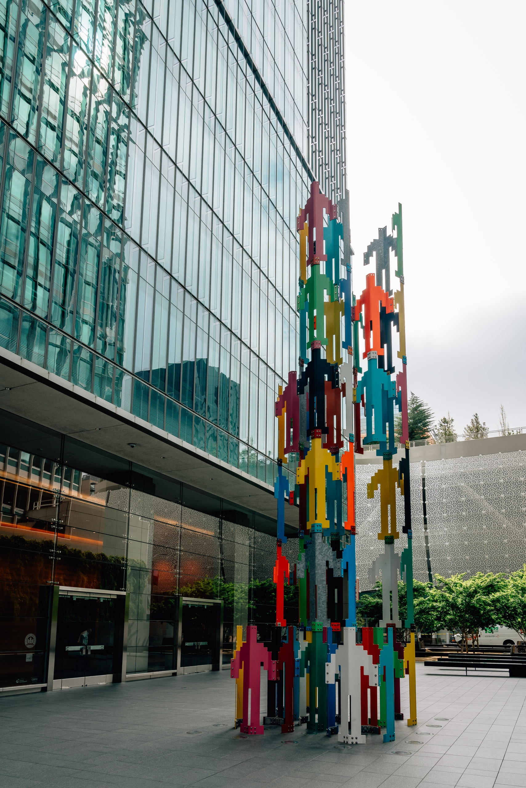 A vibrant, multicolored pixelated sculpture towers in an urban plaza beside a modern glass-fronted building under overcast skies