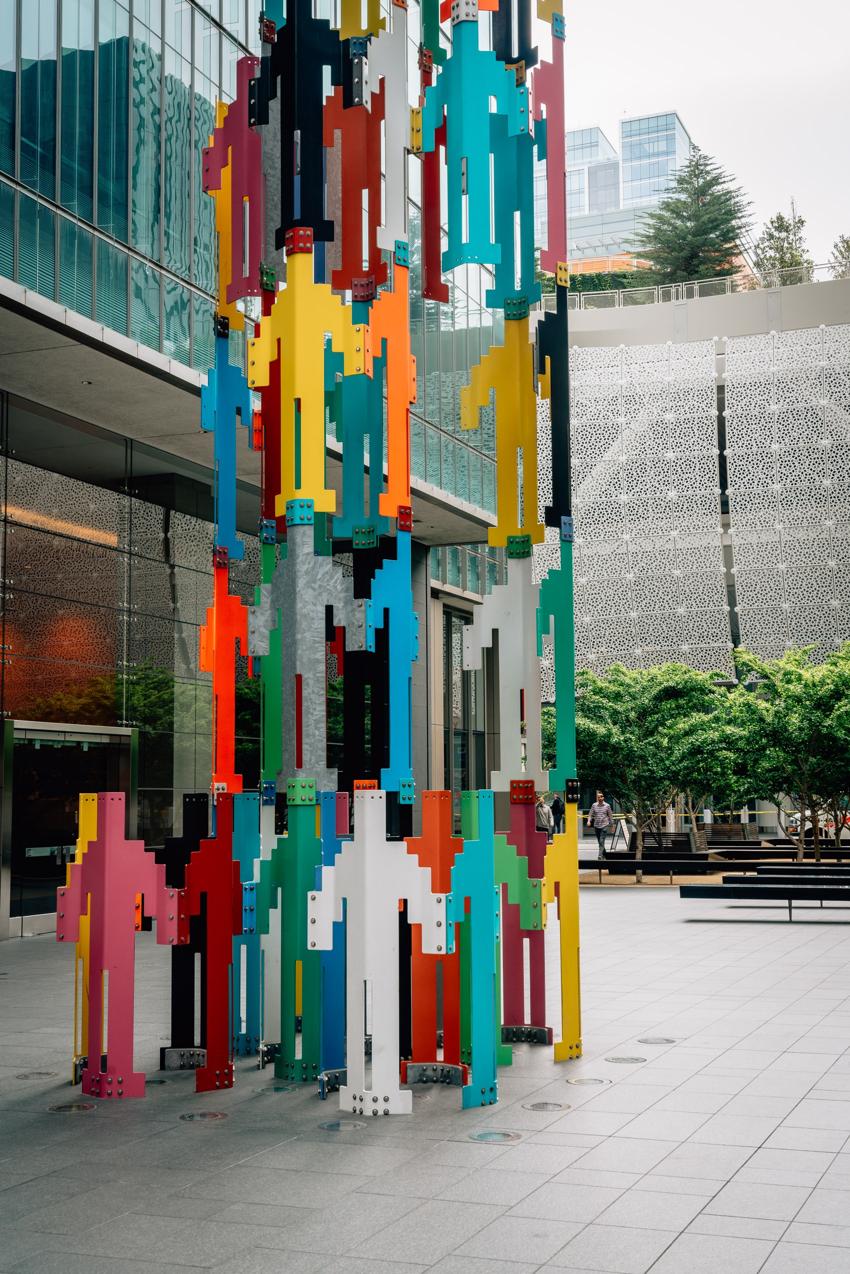 A vibrant pixelated tower sculpture in bright primary colors stands in an urban plaza surrounded by modern glass buildings and concrete architecture.