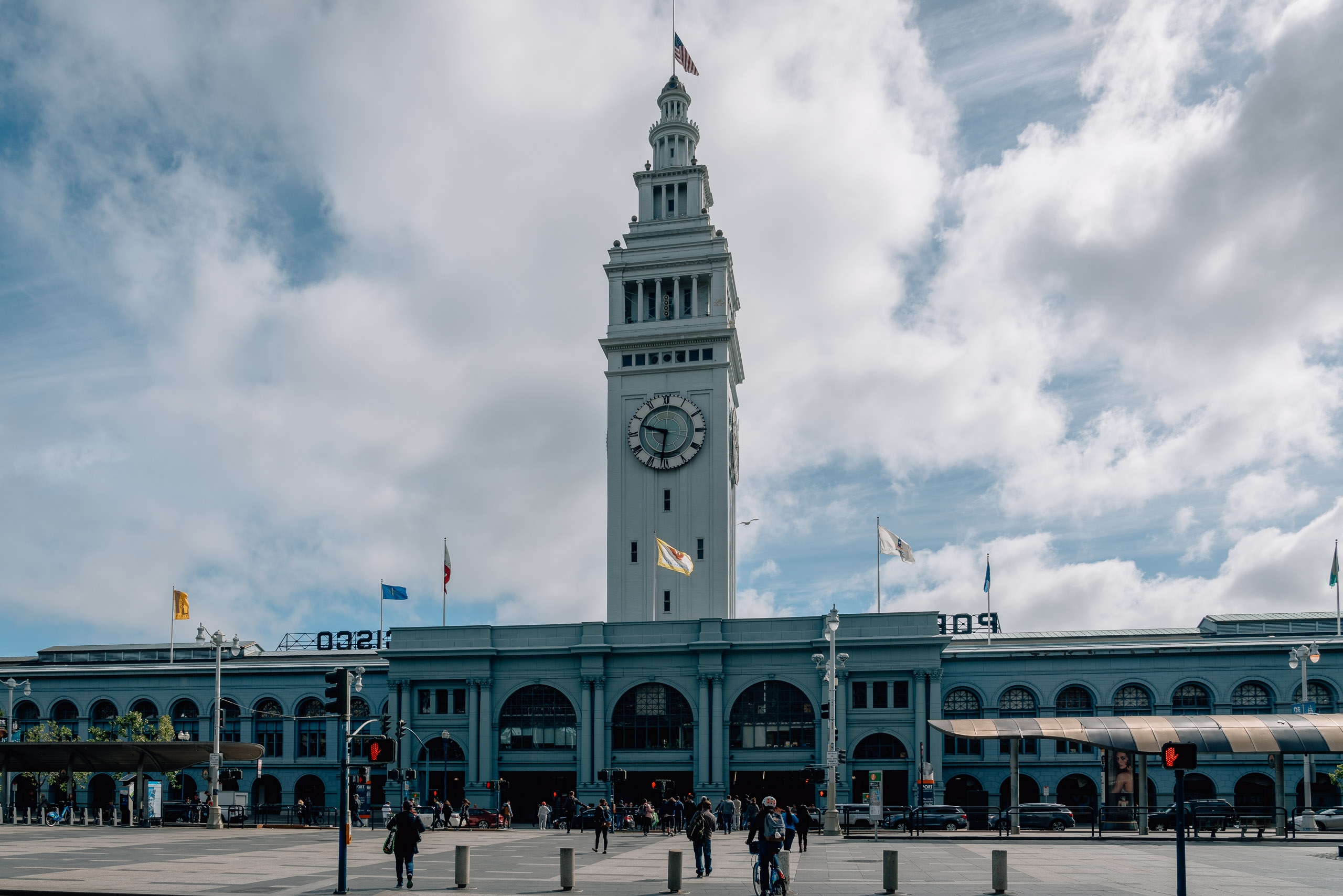 The Ferry Building's iconic clock tower rises above its distinctive turquoise arcaded structure, with an American flag at its peak against a partly cloudy sky and plaza below