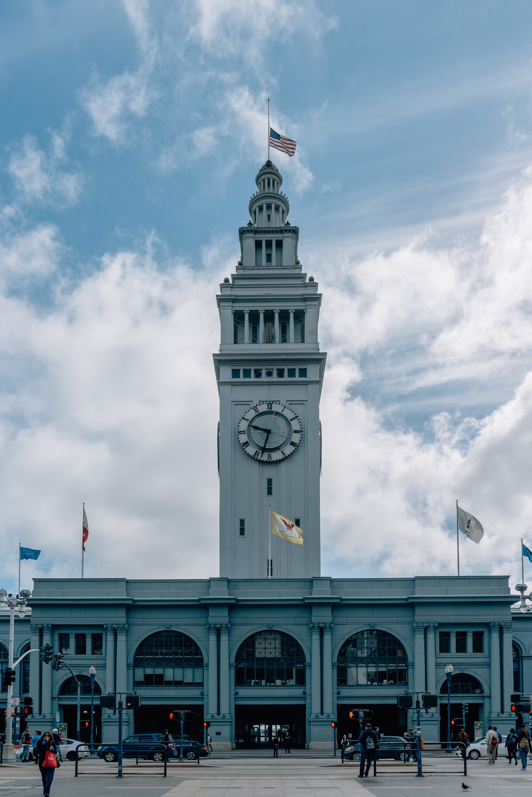 The Ferry Building Marketplace stands prominently with its distinctive clock tower rising against a partly cloudy sky, American flag flying from the spire and colorful banners draped on the facade