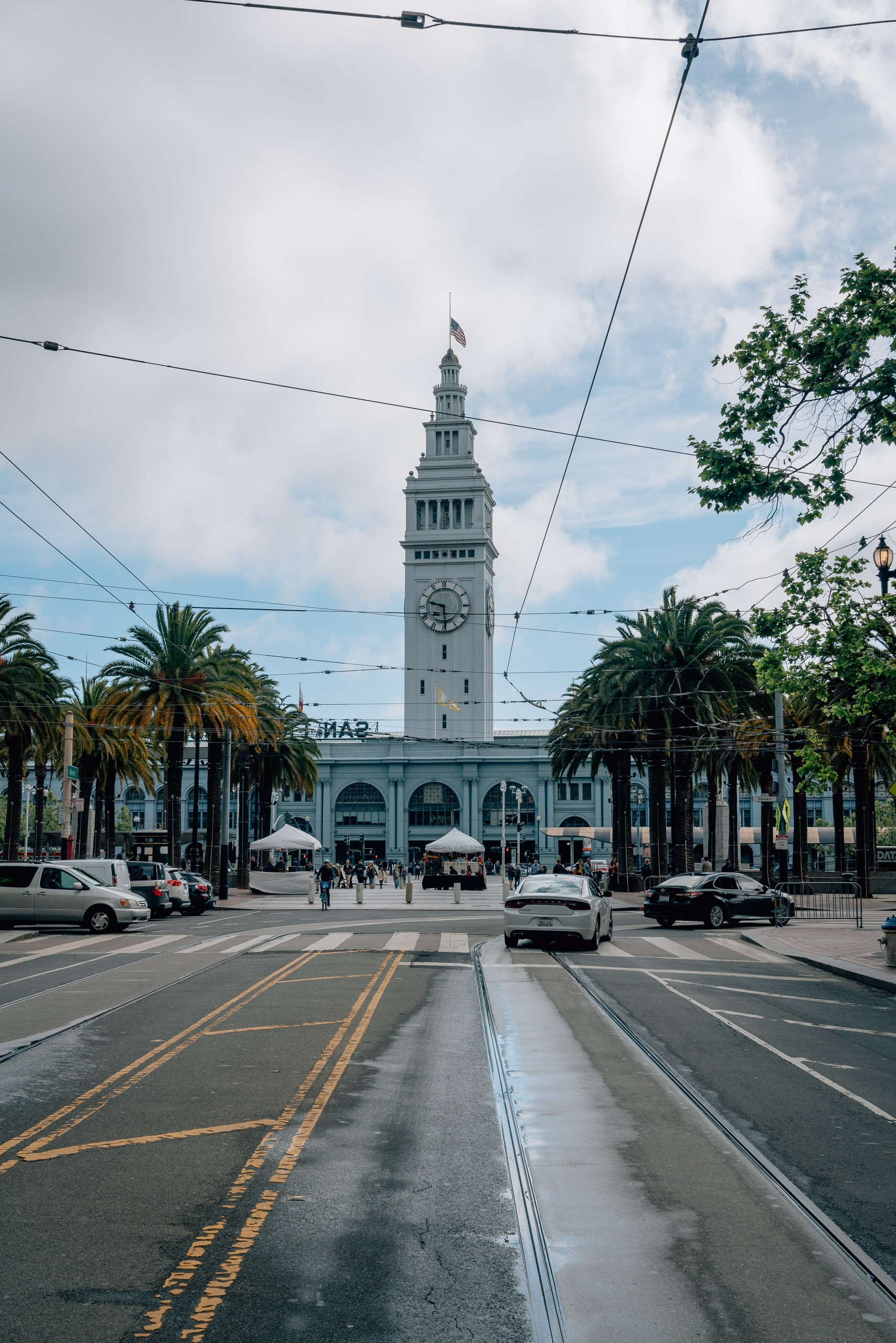 Ferry Building Marketplace stands prominently at the end of a tree-lined street with overhead trolley wires and light traffic under partly cloudy skies