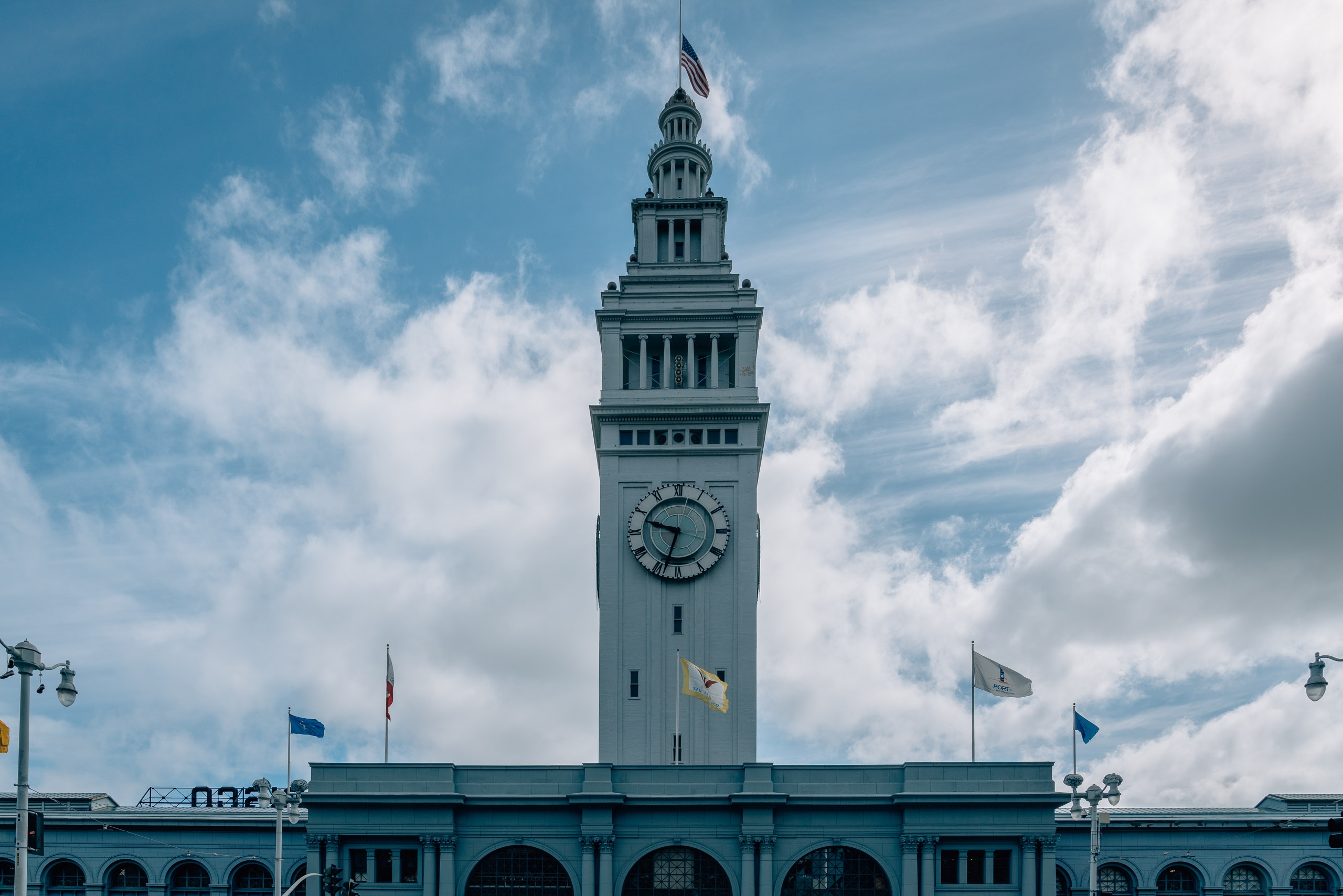 The Ferry Building's iconic clock tower rises against a bright blue sky with white clouds, its pale green structure and prominent clock face dominating the composition above the building's arched base with flags.