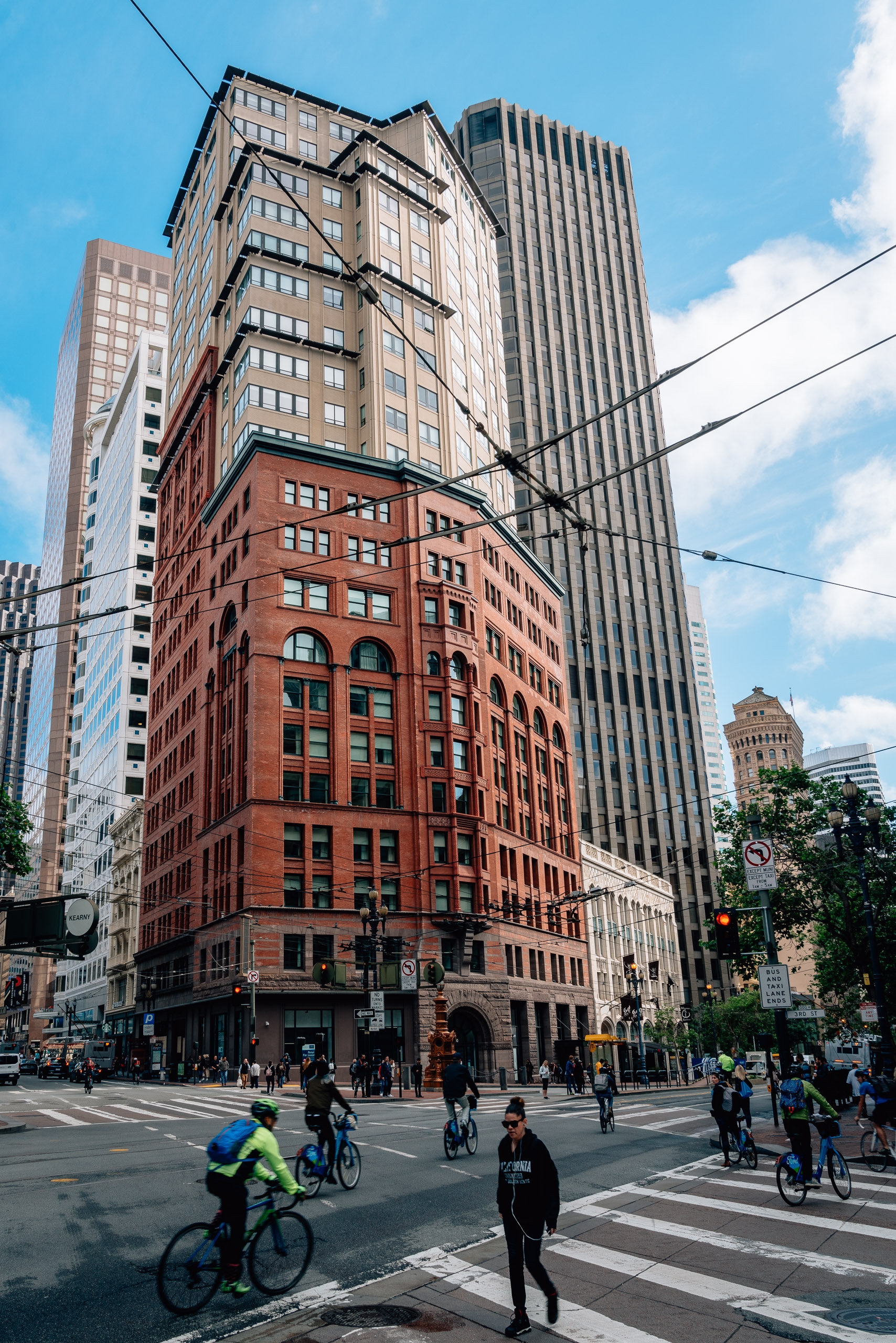 Urban intersection showcasing a red brick historic building surrounded by modern glass and steel skyscrapers, with cyclists and pedestrians crossing striped crosswalks beneath clear blue sky